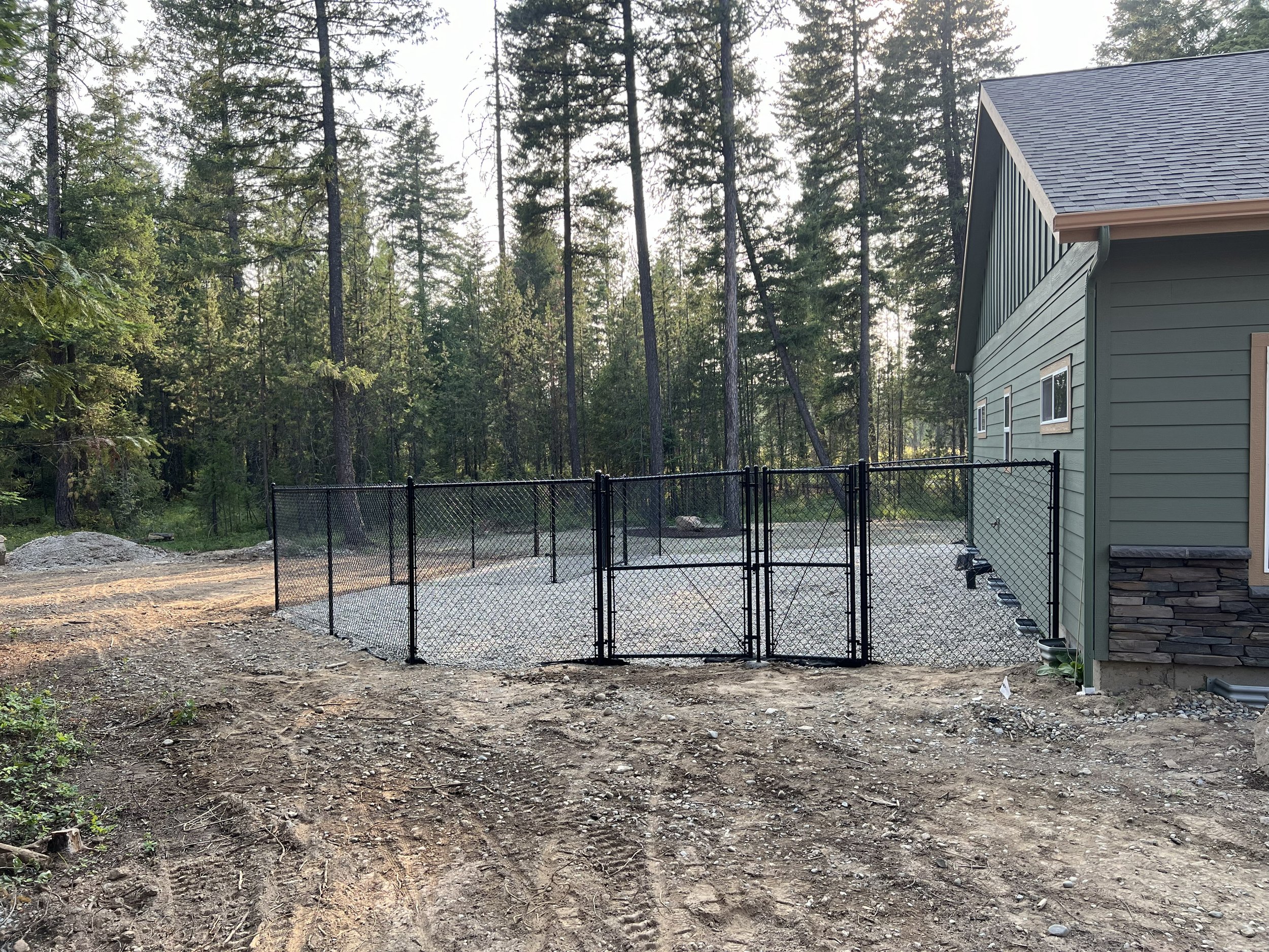 Image of a fenced backyard area with a gravel surface, attached to a green house with siding and a stone foundation, surrounded by trees.
