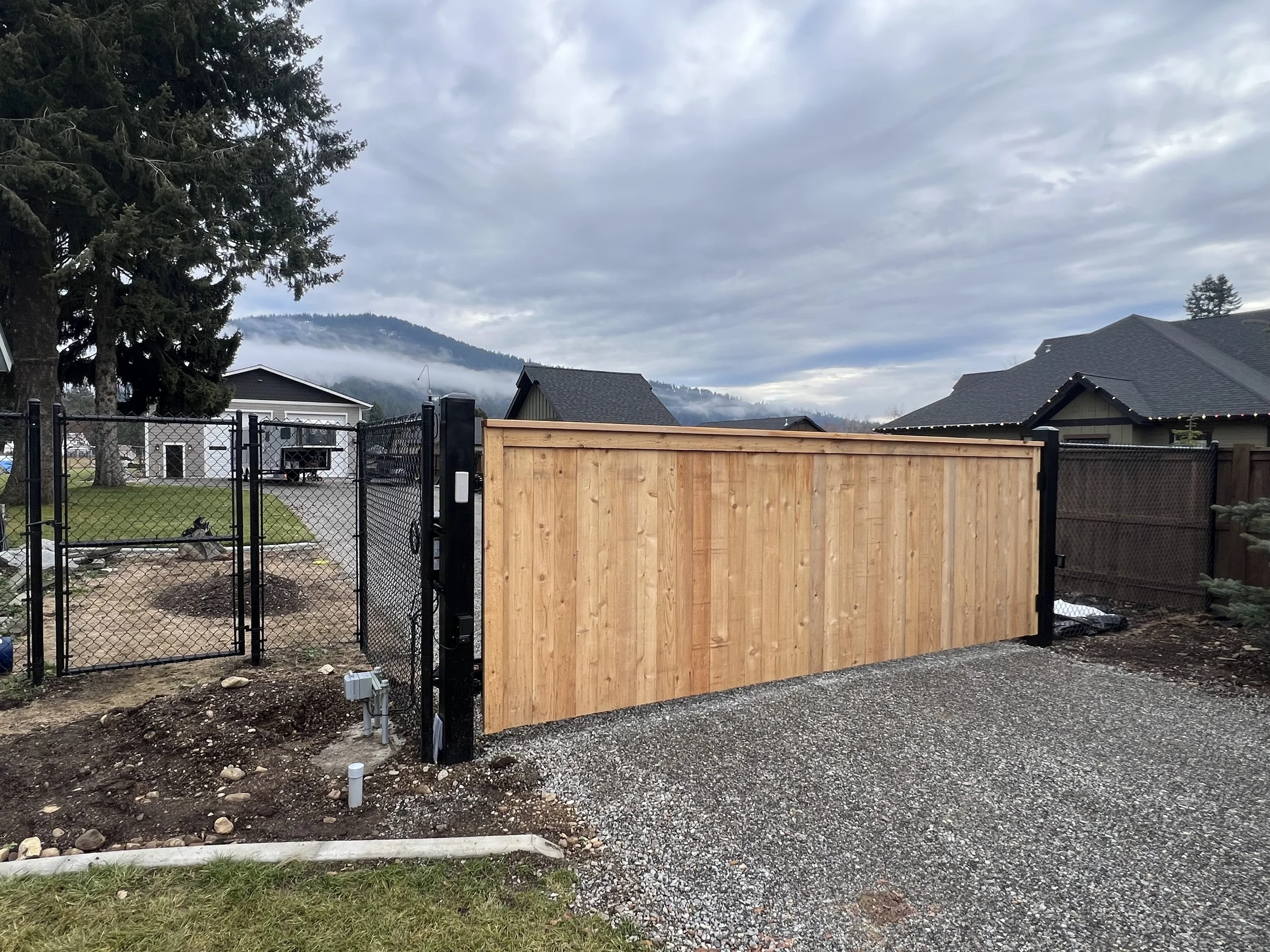A backyard with a black chain-link fence, a new wooden sliding gate, and a gravel driveway, with houses and mountains in the background under a cloudy sky.