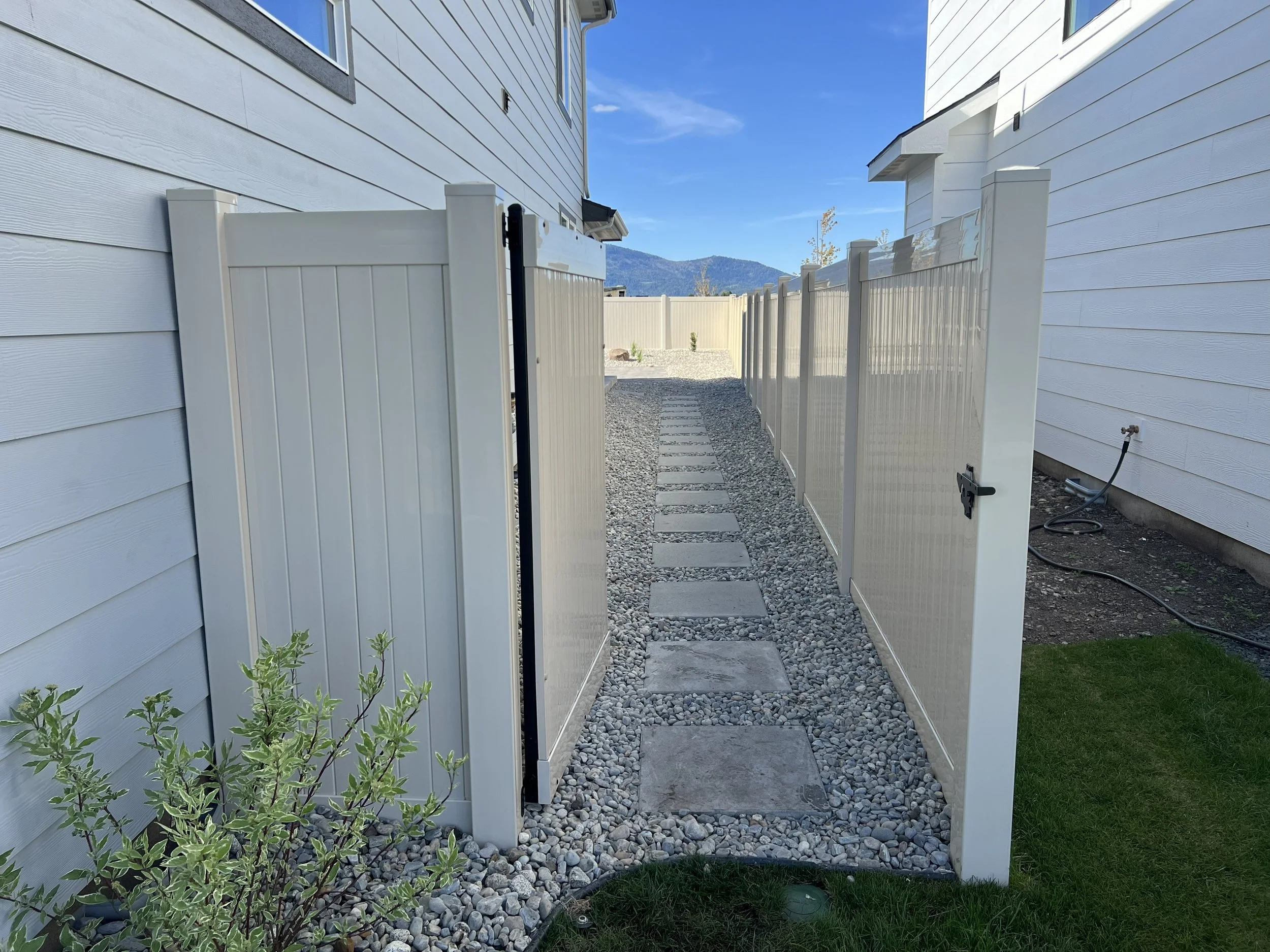 A narrow backyard pathway with small square stepping stones on gravel, flanked by white privacy fences, leading to a distant view of mountains under a blue sky.