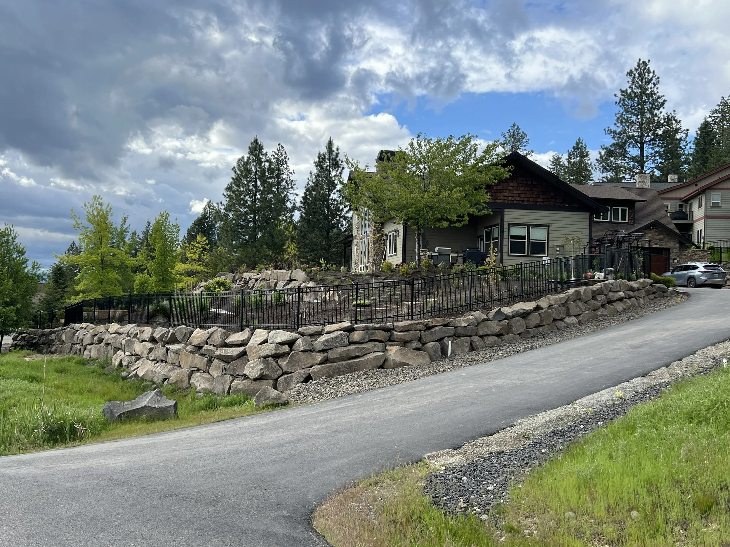 A residential house on a hill with a driveway leading up to it, surrounded by rocks and greenery, with trees and cloudy sky in the background.