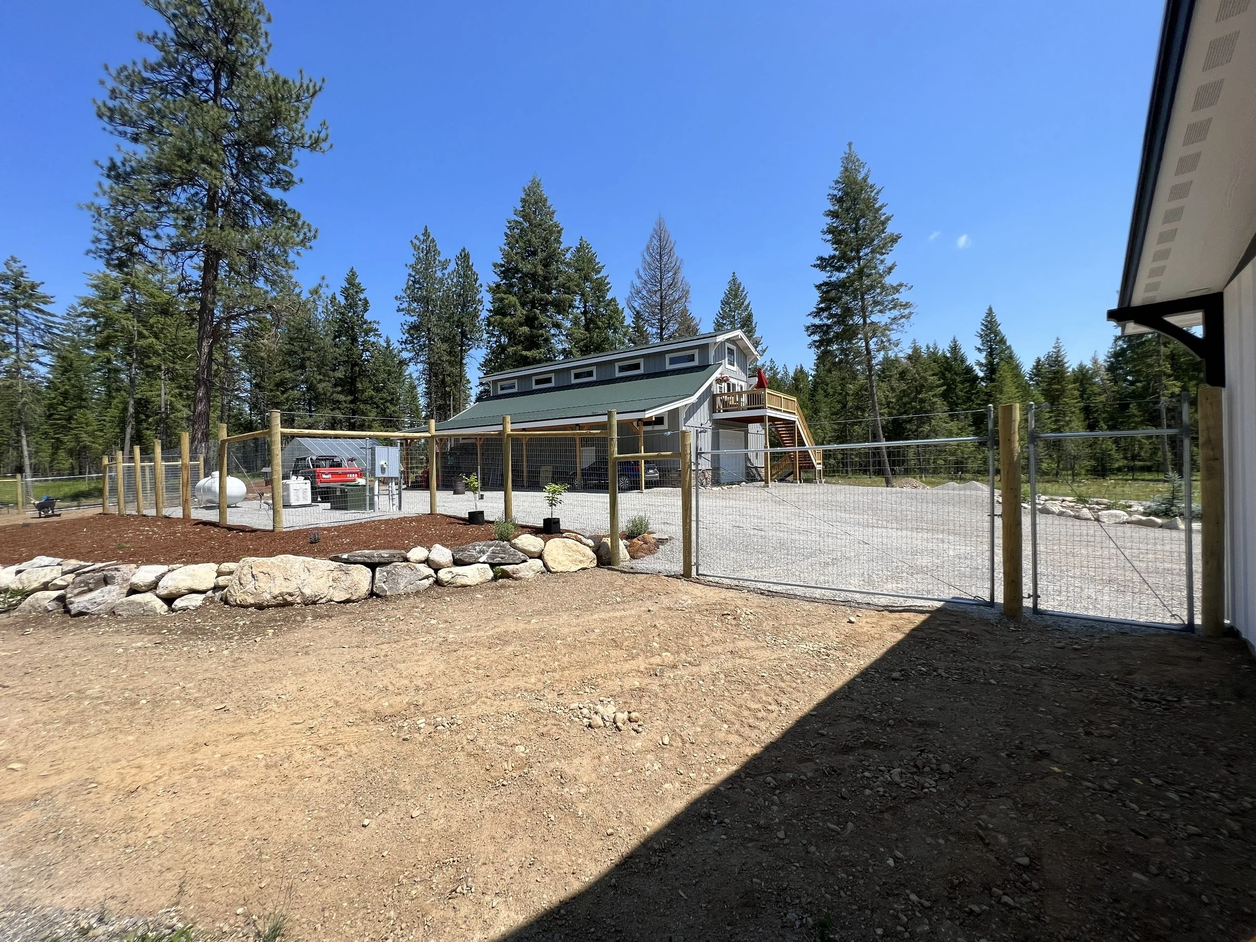 View of a backyard with a gravel driveway, a wooden fence, a two-story house with a deck, and tall pine trees under a clear blue sky.