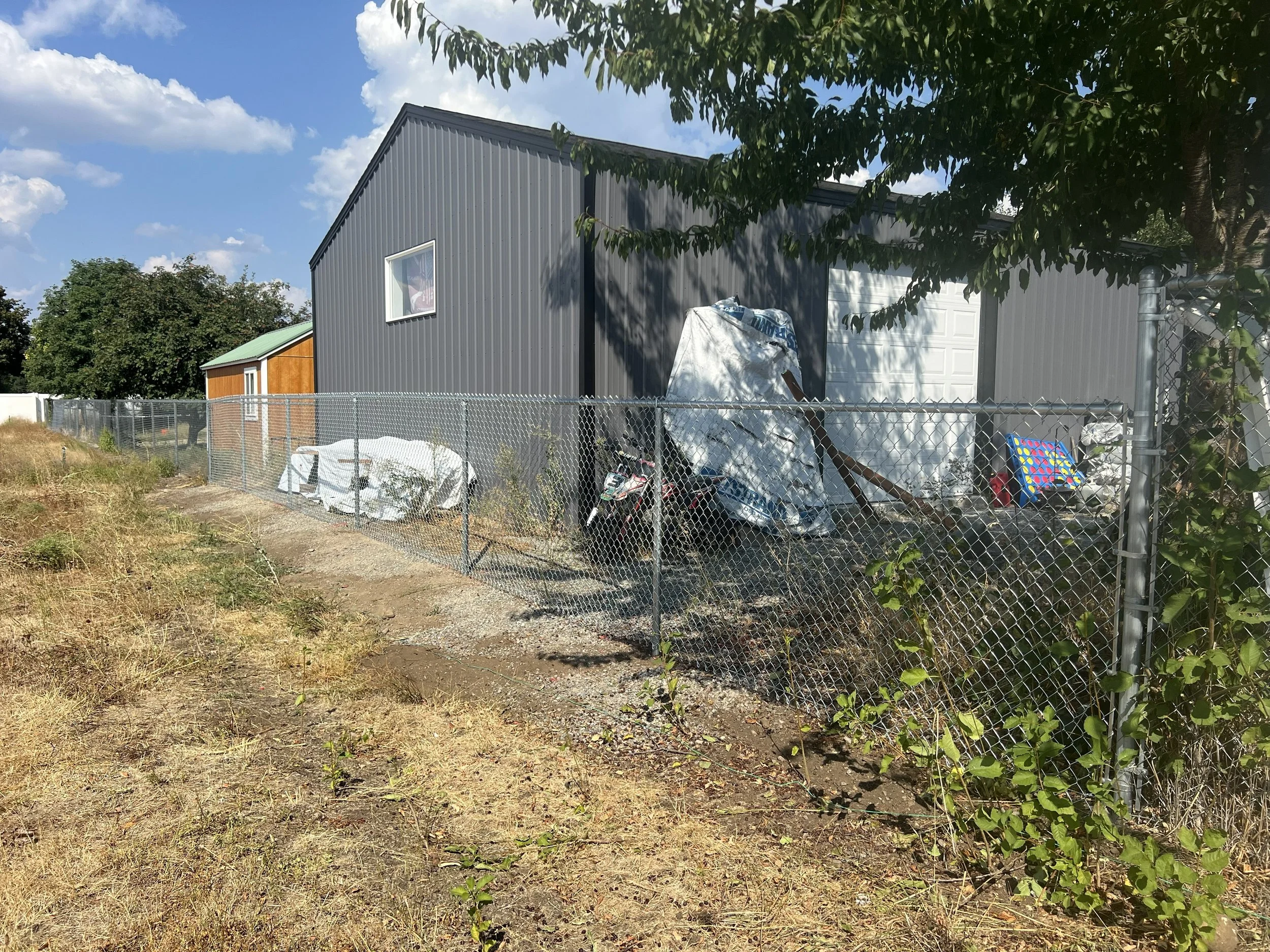 A backyard with a gray metal building, a chain-link fence, a covered motorcycle, and some outdoor items, with a grassy and dirt area in the foreground and a tree on the right side.
