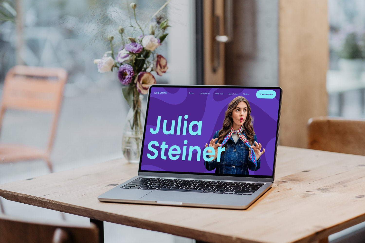Ein Laptop auf einem Holztisch zeigt eine Webseite mit dem Namen Julia Steiner, auf denen eine Frau mit langem braunem Haar und einem bunten Halstuch zu sehen ist.