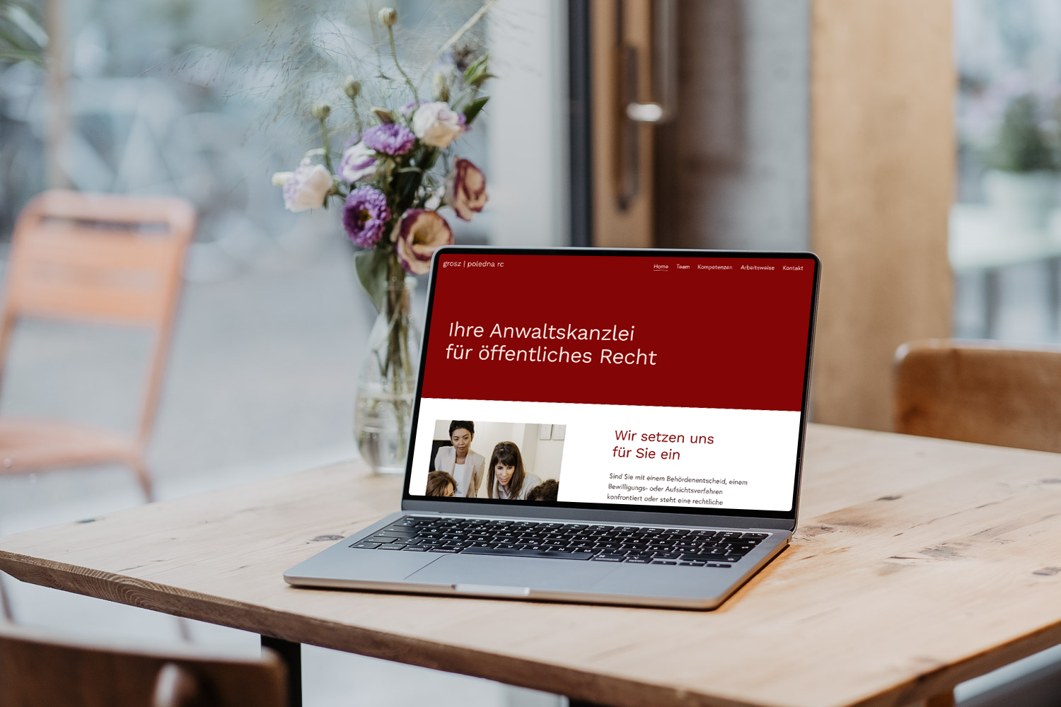 Laptop auf einem Holztisch mit einer Anwaltskanzlei Website, neben einem Blumenstrauß in einer Vase, im Hintergrund ein stimmungsvoller Raum mit Stühlen.