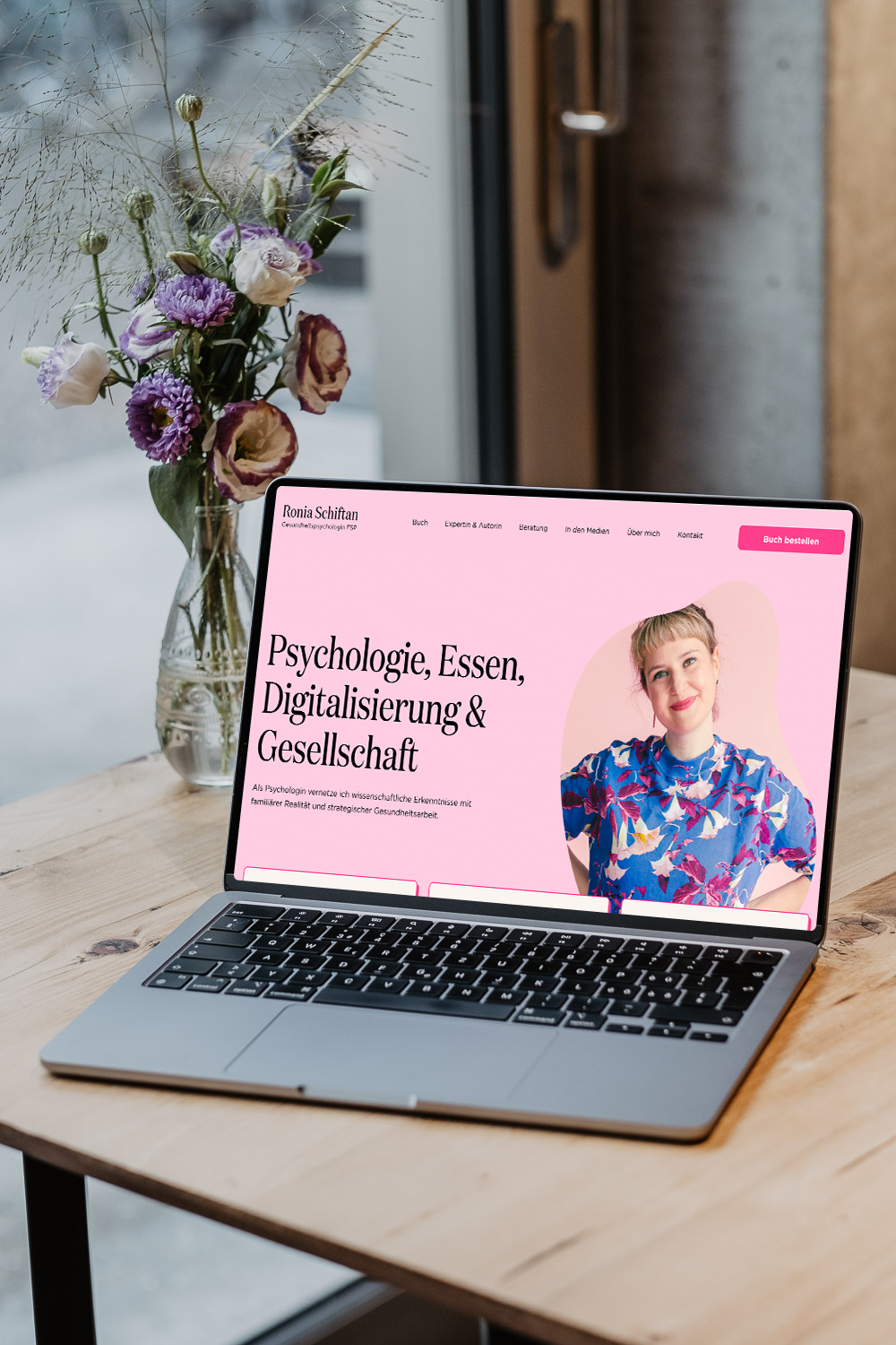 Laptop auf einem Holztisch mit einer Website über Psychologie, Essen, Digitalisierung und Gesellschaft, daneben eine Vase mit Blumen.