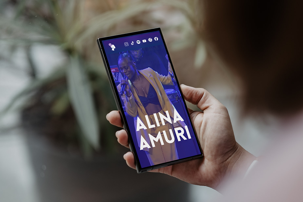 Hand hält Smartphone mit Bild einer Frau, die auf einer Bühne singt, mit Text 'ALINA AMURI' auf dem Bildschirm.