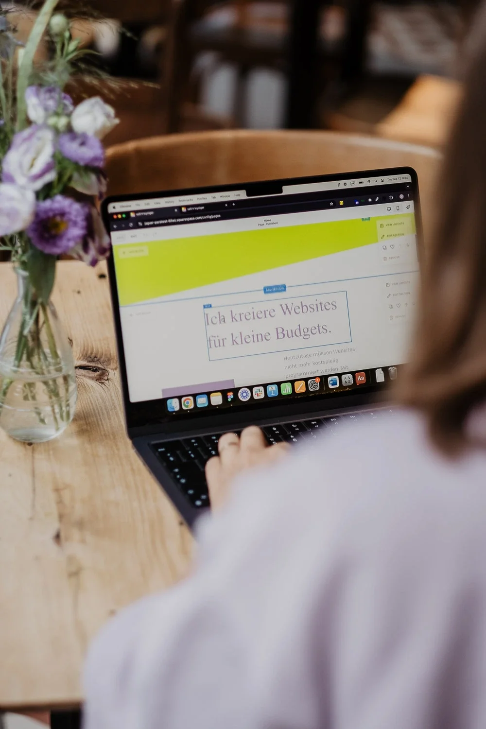 Ein Laptop, auf dem eine Webseite mit dem Text 'Ich kreire Websites für kleine Budgets' geöffnet ist, befindet sich auf einem Holztisch neben einem Glas mit Blumen.