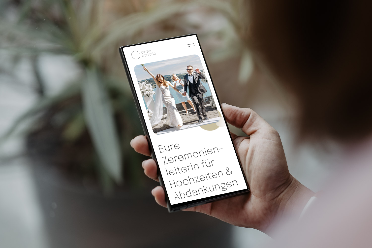 Hand hält ein Smartphone mit einer Website für Hochzeitszeremonien, die ein Bild von einem Hochzeitspaar zeigt und den Text 'Eure Zeremonienleiterin für Hochzeiten & Abdankungen' enthält.