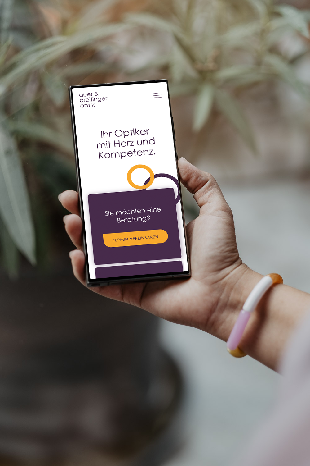 Hand hält ein Smartphone mit einer Website für eine Optikerfirma, auf der steht: 'Ihr Optiker mit Herz und Kompetenz. Möchten Sie eine Beratung? Termin vereinbaren'.
