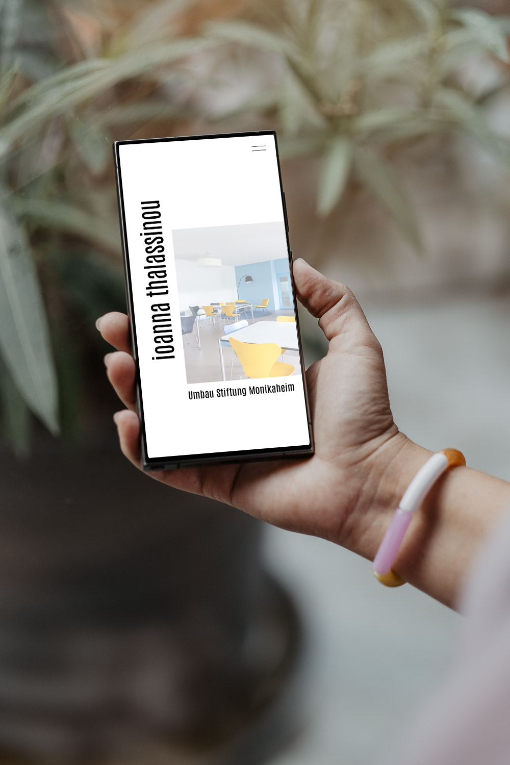 Hand hält Smartphone mit einer Präsentation über den Umbau der Stiftung Monikaheims, im Hintergrund Pflanzen.