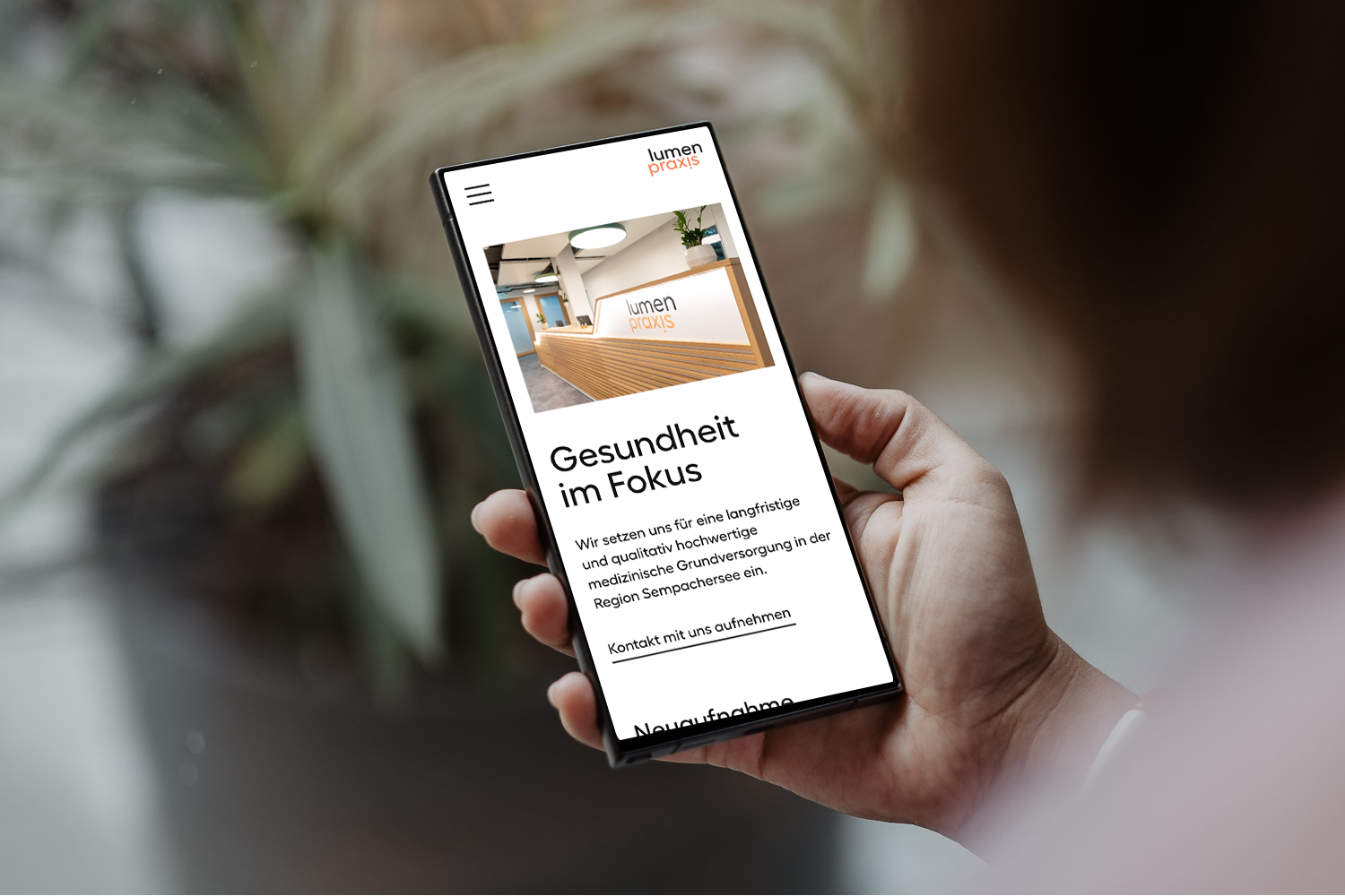 Hand hält ein Smartphone, das eine Webseite mit dem Titel "Gesundheit im Fokus" zeigt. Im Hintergrund sind Pflanzen sichtbar.
