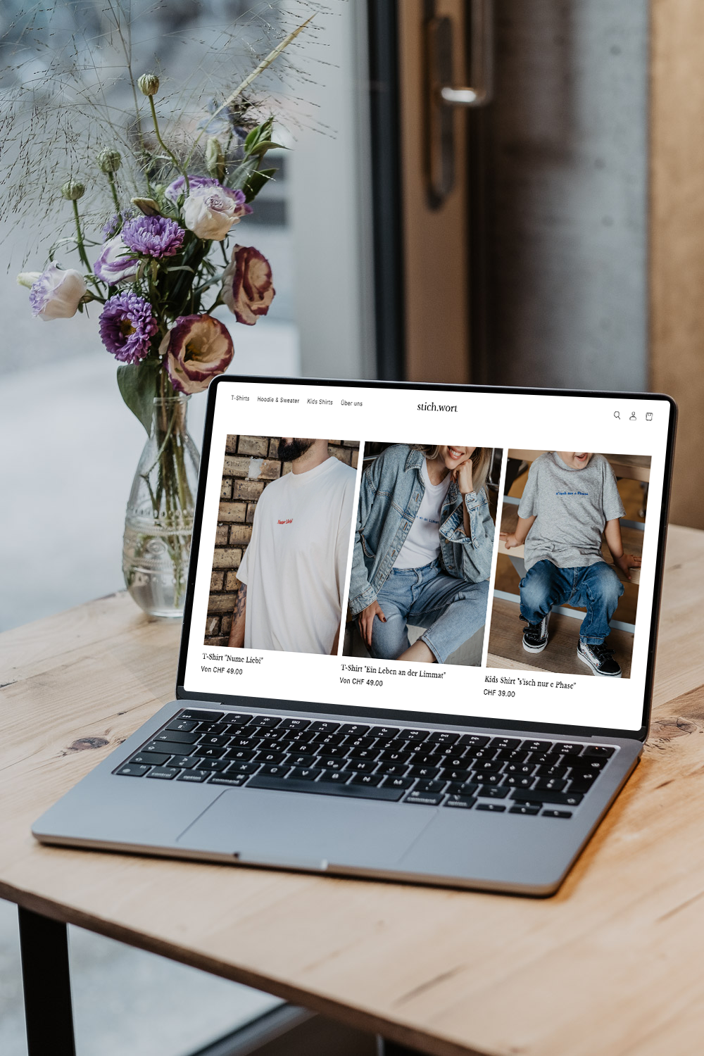 Laptop auf einem Holztisch mit einem Blumenstrauß in einer Glasvase im Hintergrund. Der Bildschirm zeigt einen Online-Shop für T-Shirts mit Bildern von Menschen in verschiedenen Outfits, Preise und Beschreibungen in Deutsch.