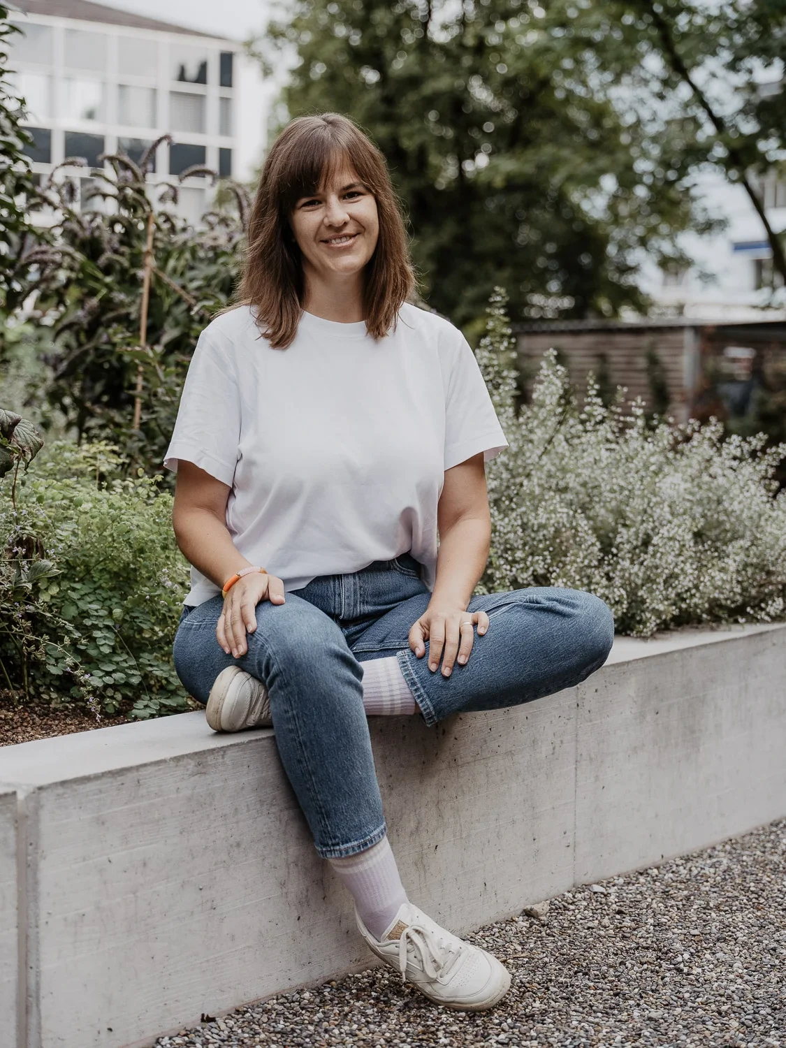 Gaby Jenni sitzt auf einem erhöhten Betonrand vor Grünpflanzen und Bäumen in einem Garten von einem Café.
