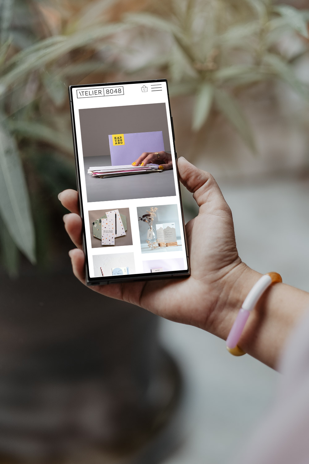 Nahaufnahme einer Hand, die ein Smartphone hält und einen Online-Shop für Design- und Papeterieartikel zeigt.