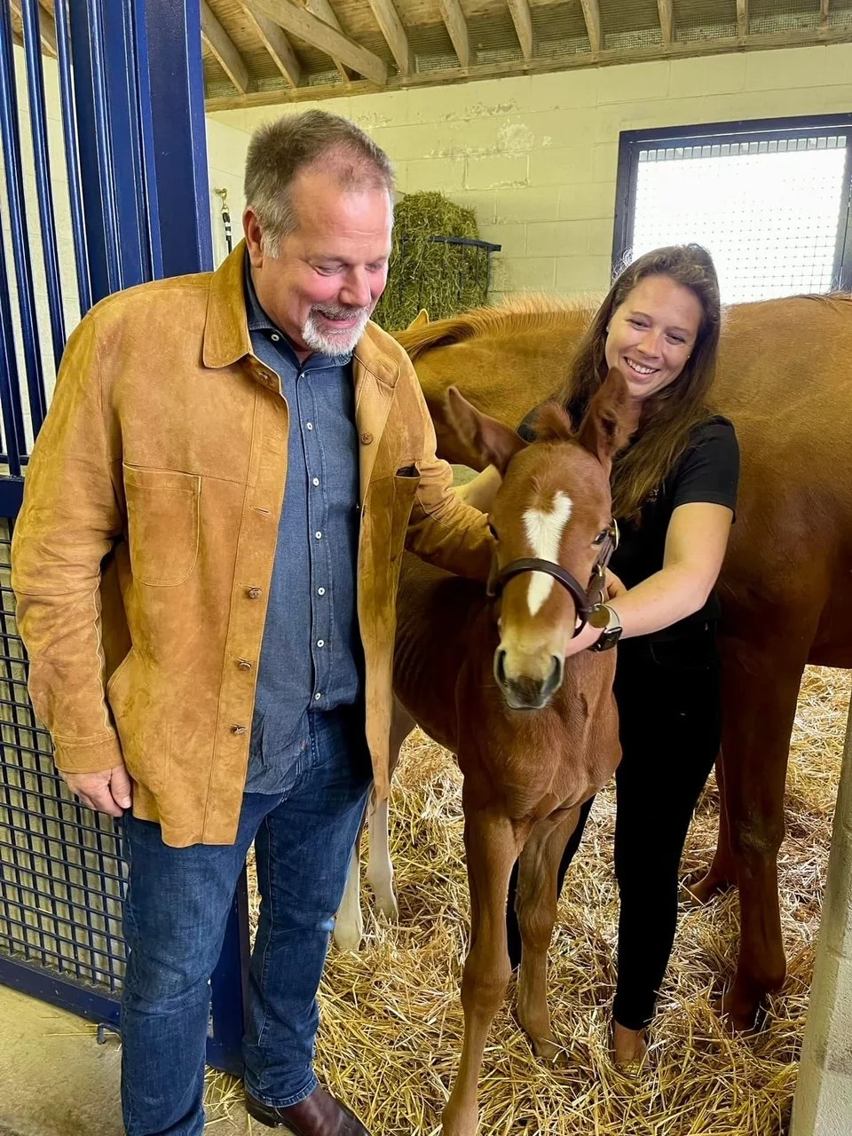A man and a woman standing with a baby horse inside a barn, smiling and petting the foal.