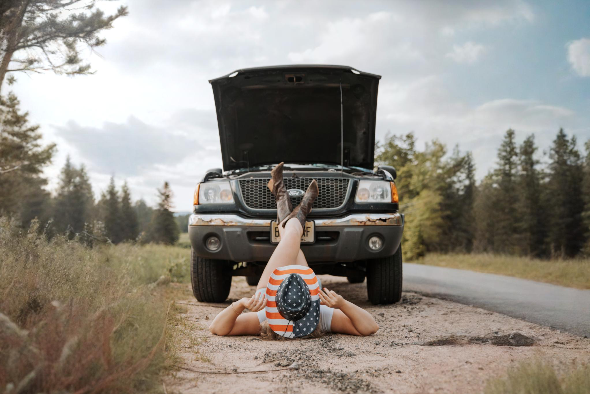 Person lying on the ground with legs up on the hood of a black SUV with an open hood, looking underneath. The person is wearing a patriotic hat and boots, and is surrounded by a rural landscape with trees and a partly cloudy sky.