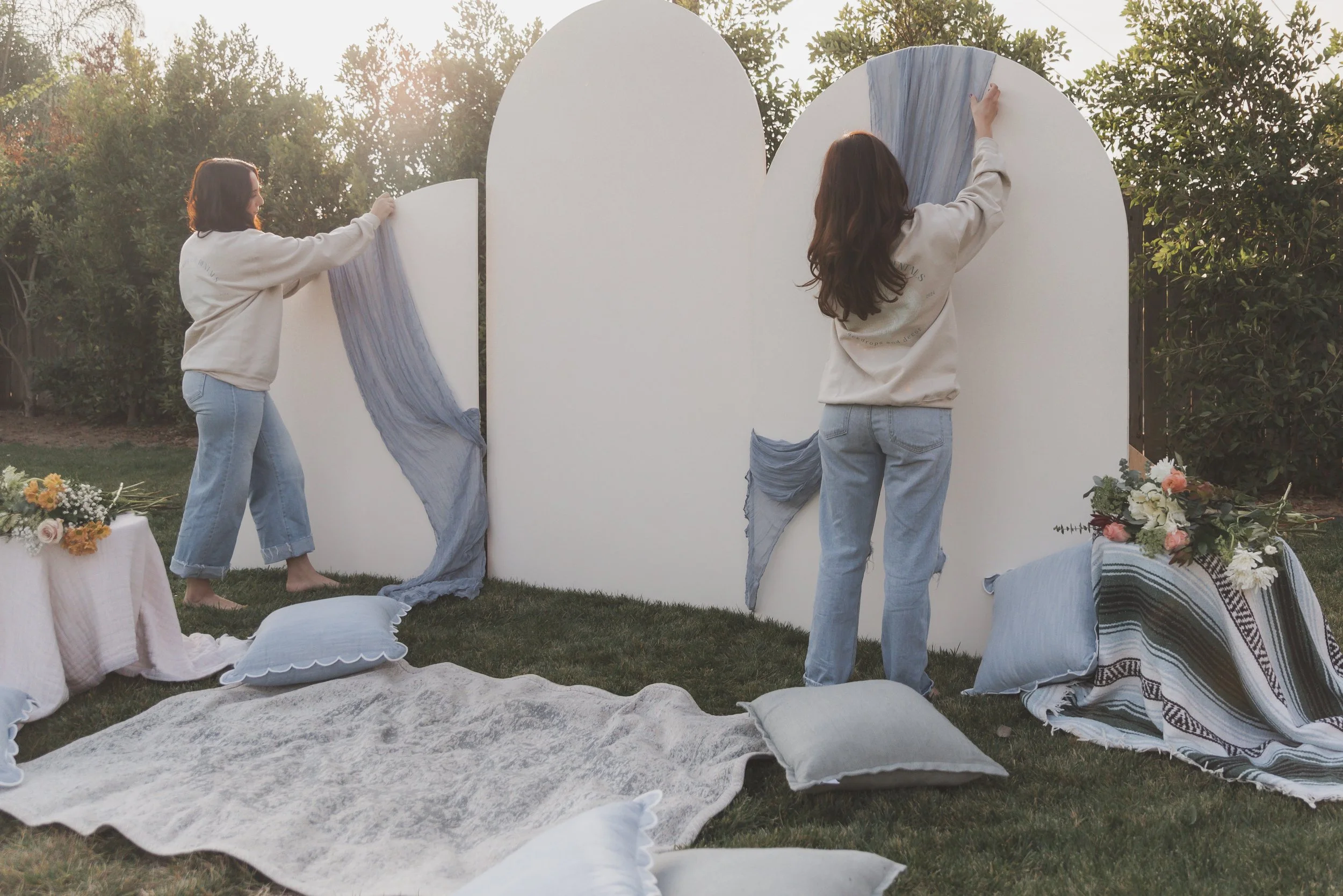 Two women setting up a fabric backdrop with blue and white fabric outdoors in a garden, with cushions and floral arrangements nearby.