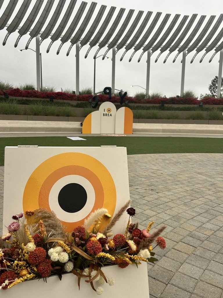A decorative outdoor installation featuring a circular target with orange, yellow, white, and black rings, surrounded by a floral arrangement with various dried flowers and grasses. In the background, a sign reads 'I Love Brea,' with a large modern s