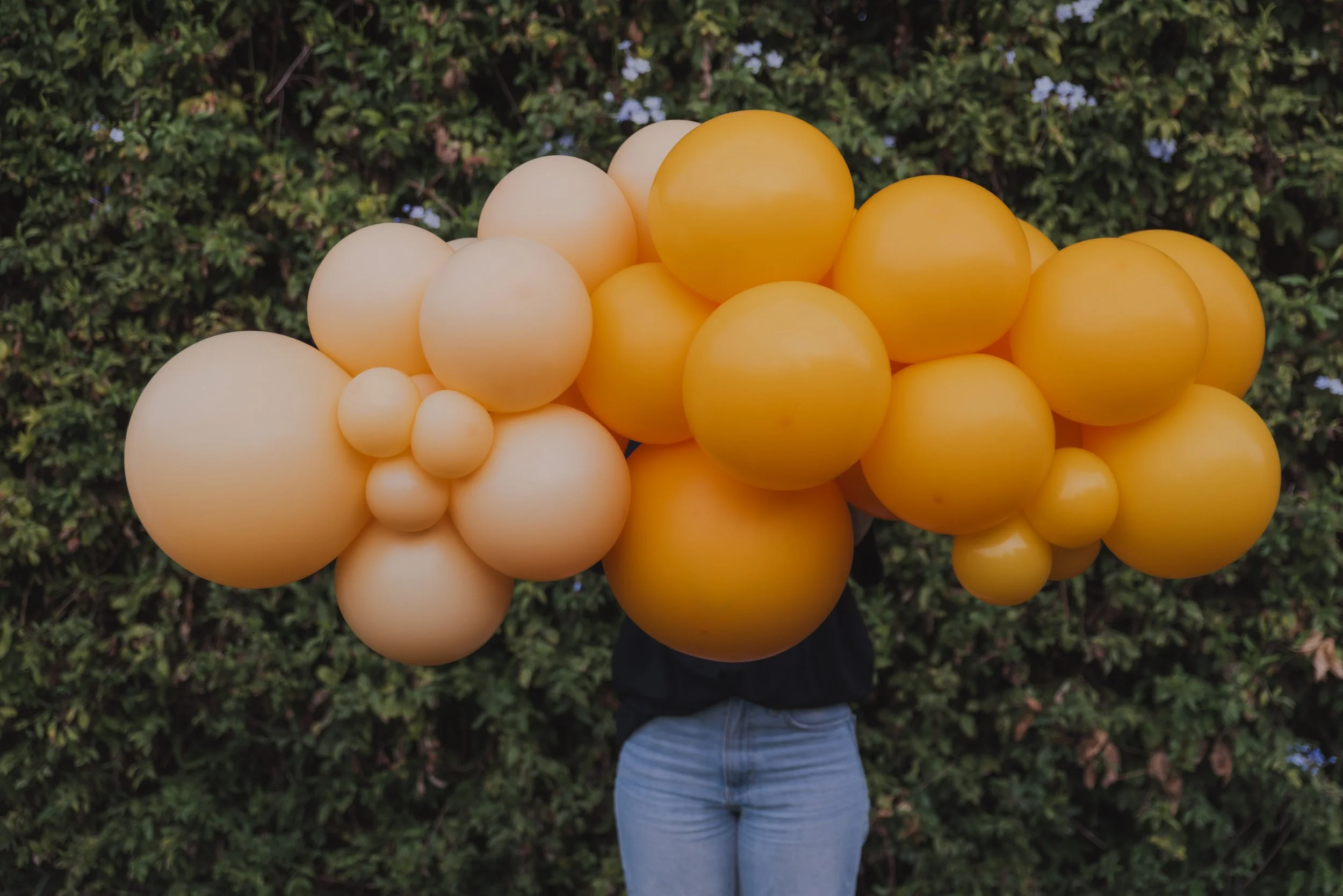 Person holding bunch of yellow and cream-colored balloons in front of greenery.