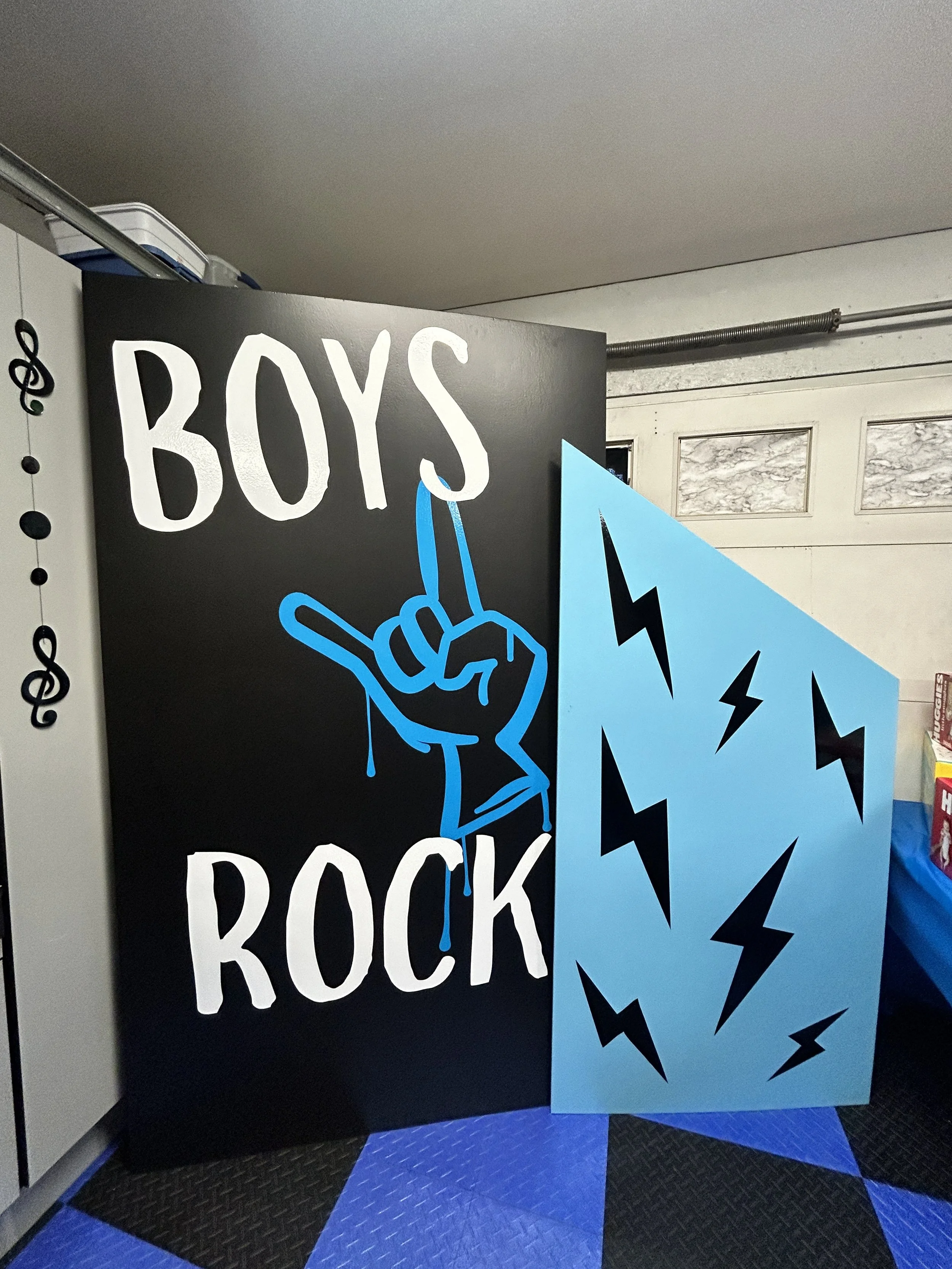 A black sign with the words 'BOYS ROCK' in white letters and a blue hand making a rock gesture outlined in blue, with some dripping paint. Next to the sign is a blue panel decorated with black lightning bolt designs.
