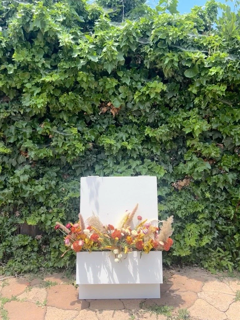 A white box with colorful dried flowers and pampas grass arranged in front of a large green leafy hedge against a partly cloudy sky.