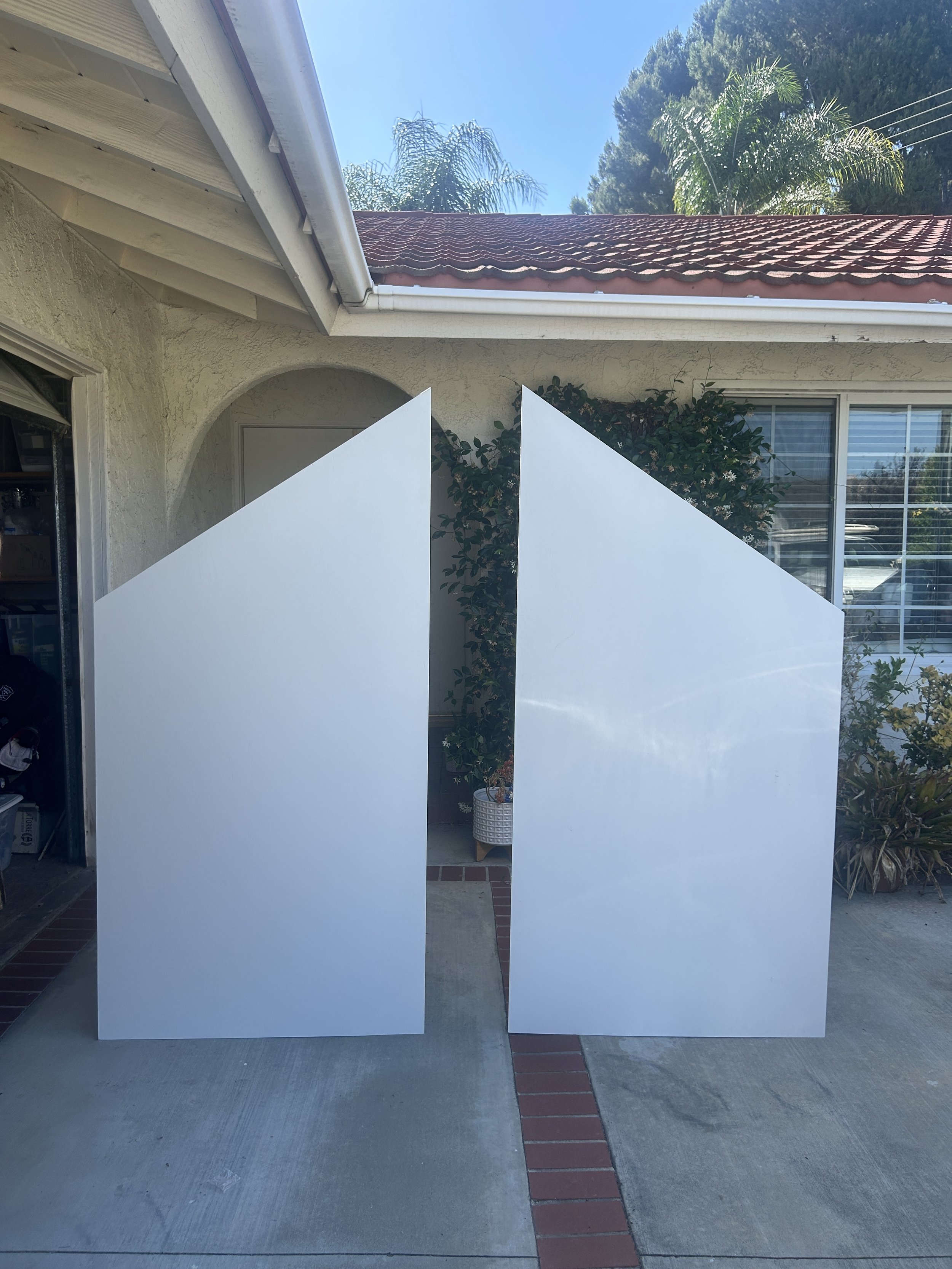 Two large white backdrops are standing outdoors on a concrete patio, with a house, window, garden, and trees in the background.