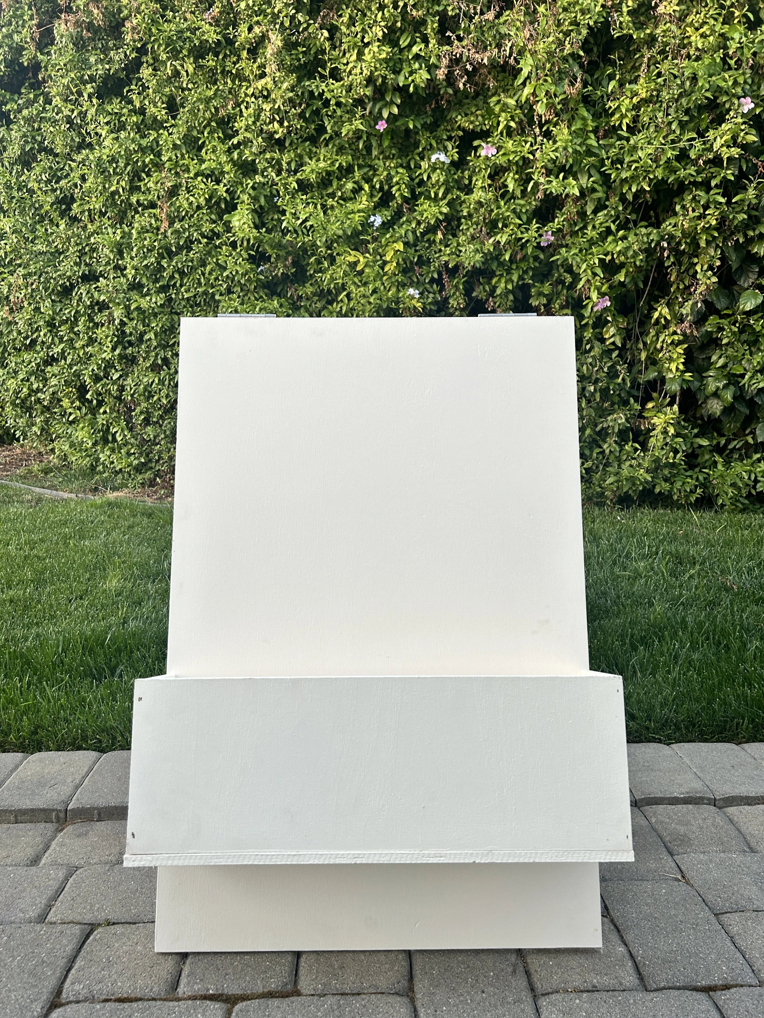 A blank white flower box sign standing on a sidewalk with green grass and a bush with purple flowers in the background.