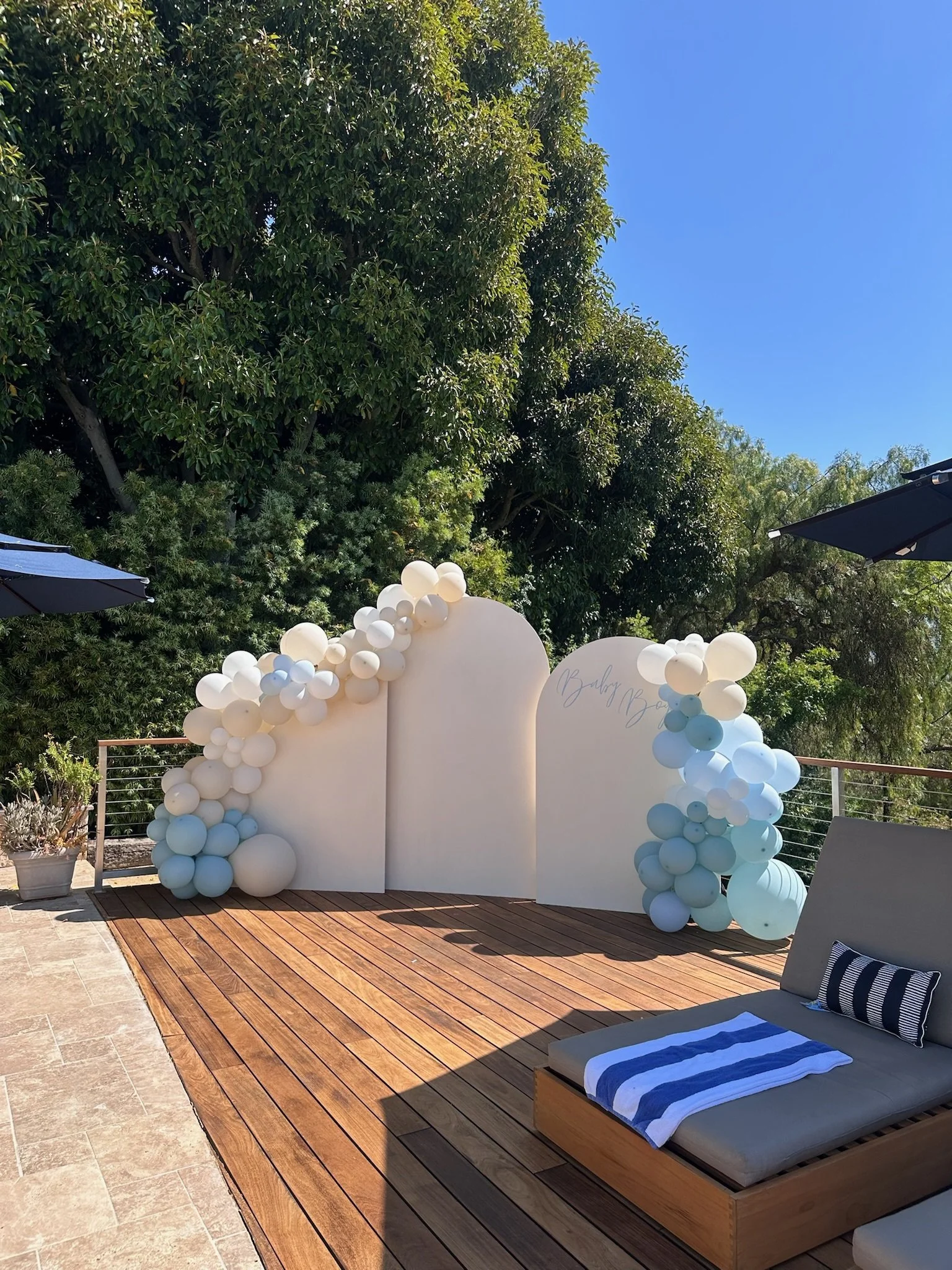 Outdoor event setup with a white balloon arch and backdrop, green trees, blue sky, a wooden deck, and a lounge chair with a striped towel.