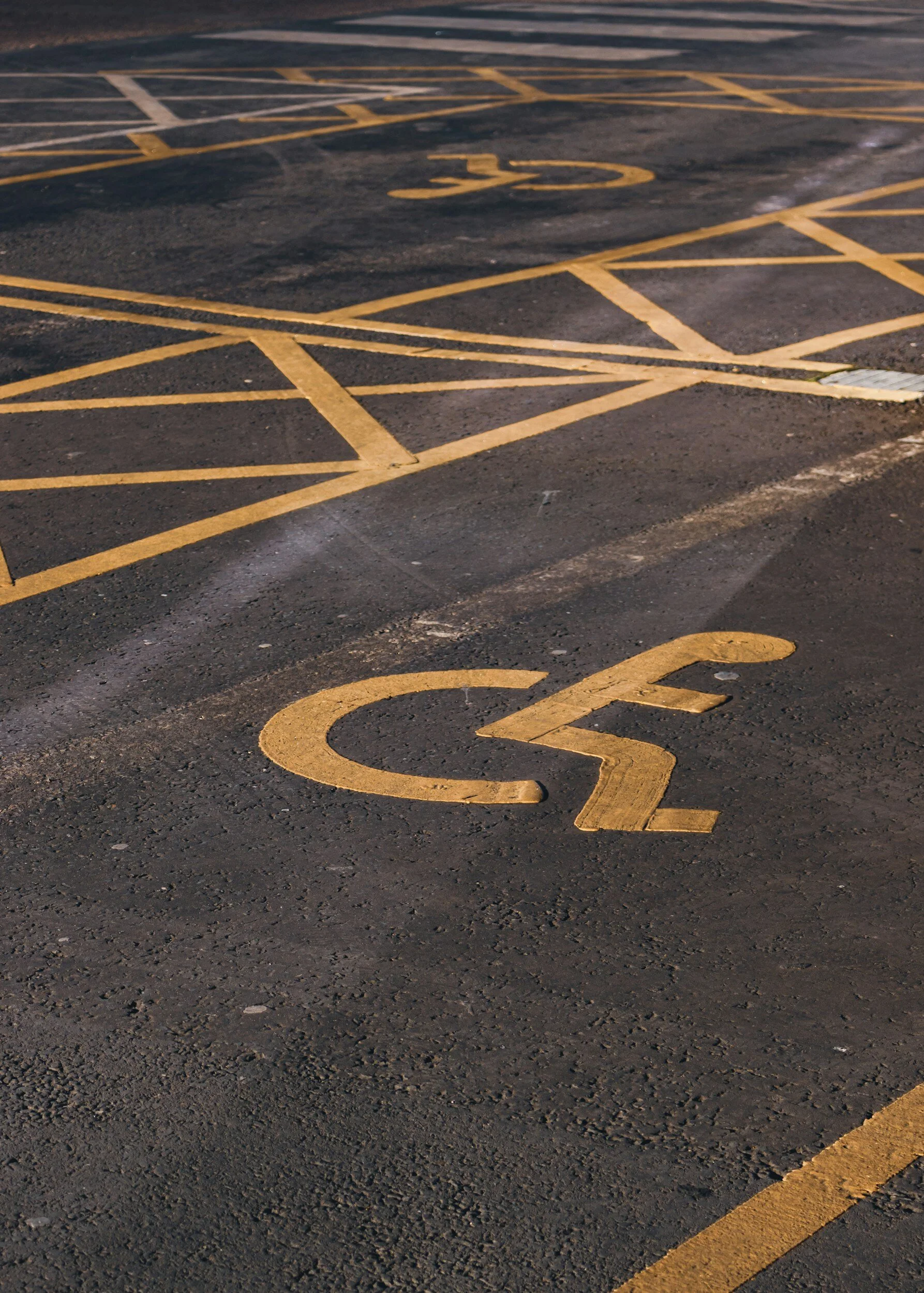 Yellow disabled parking space symbol painted on black asphalt, with additional yellow markings visible in the background.