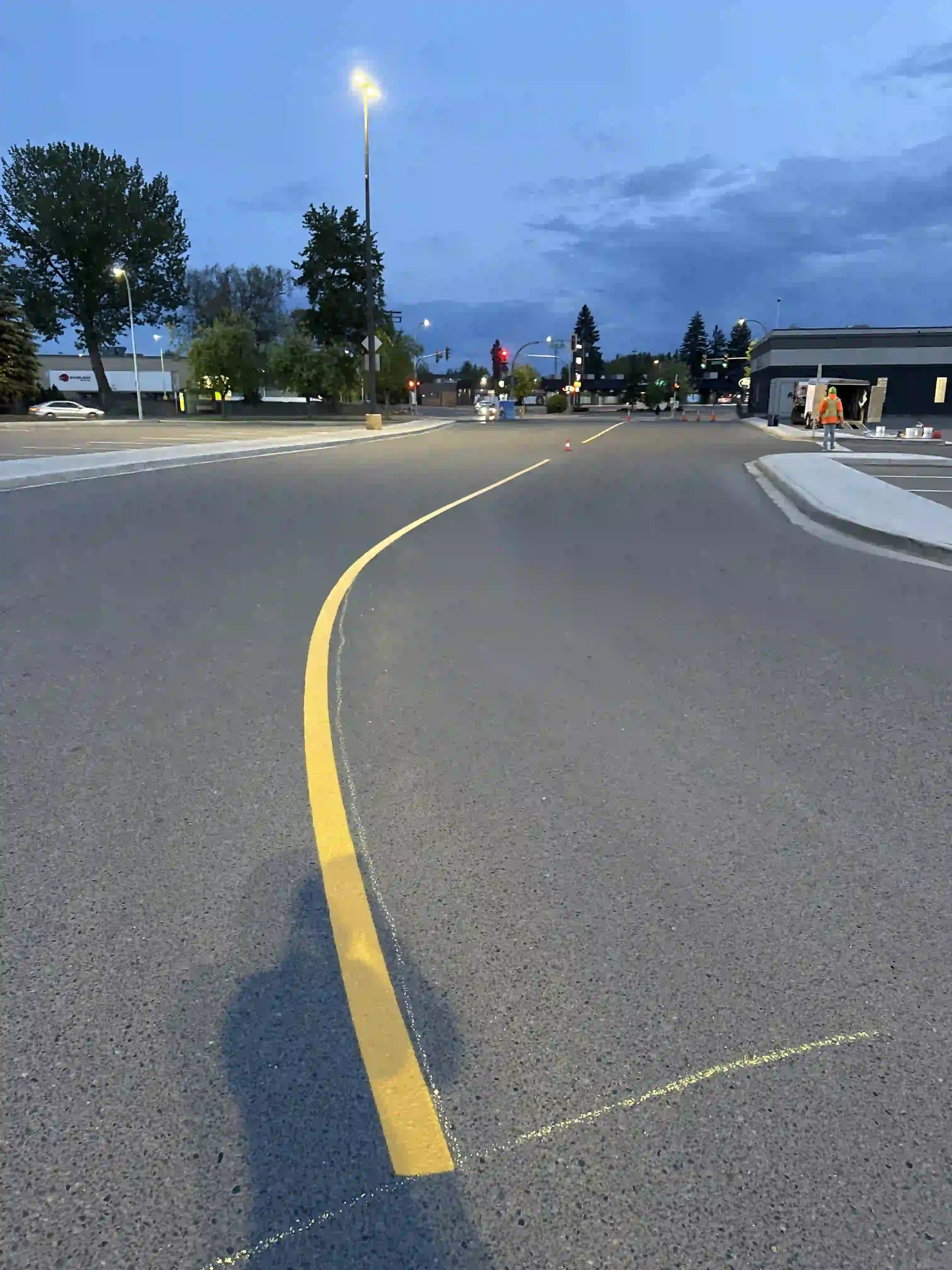 A parking lot during dawn or dusk with construction workers setting up cones, street lights illuminating the area, and a building on the right side.