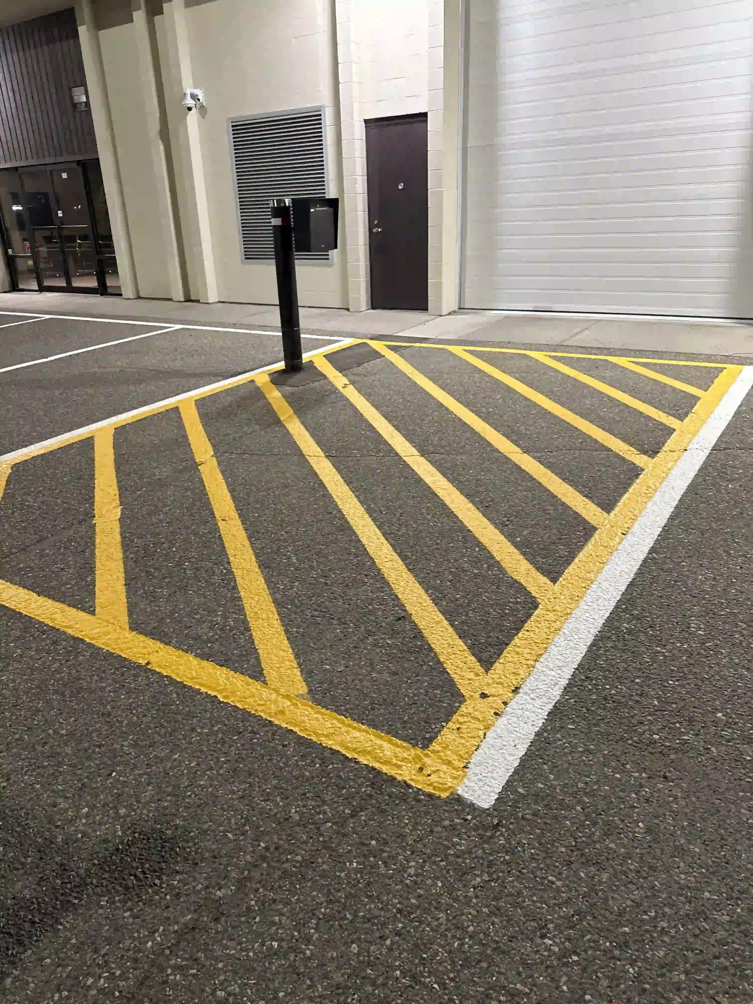 Empty parking lot with yellow-striped no-parking zone and a security camera mounted on the wall near a door. The background shows a building with a white roll-up door and a metal door.