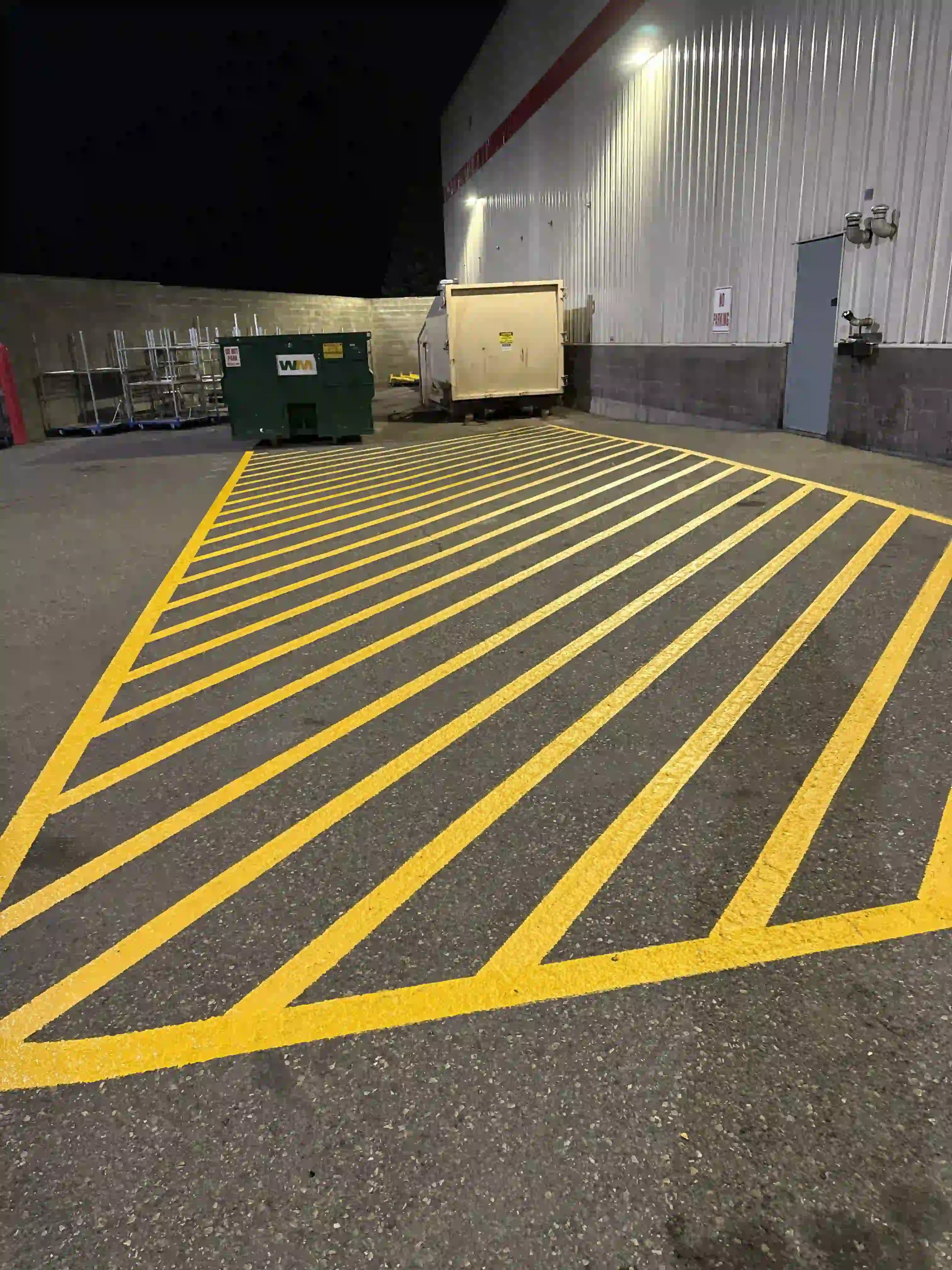 Nighttime view of a parking lot with yellow-striped warning zone markings on the pavement, a green dumpster, a white enclosed trailer, and a gray door on a corrugated metal building wall.