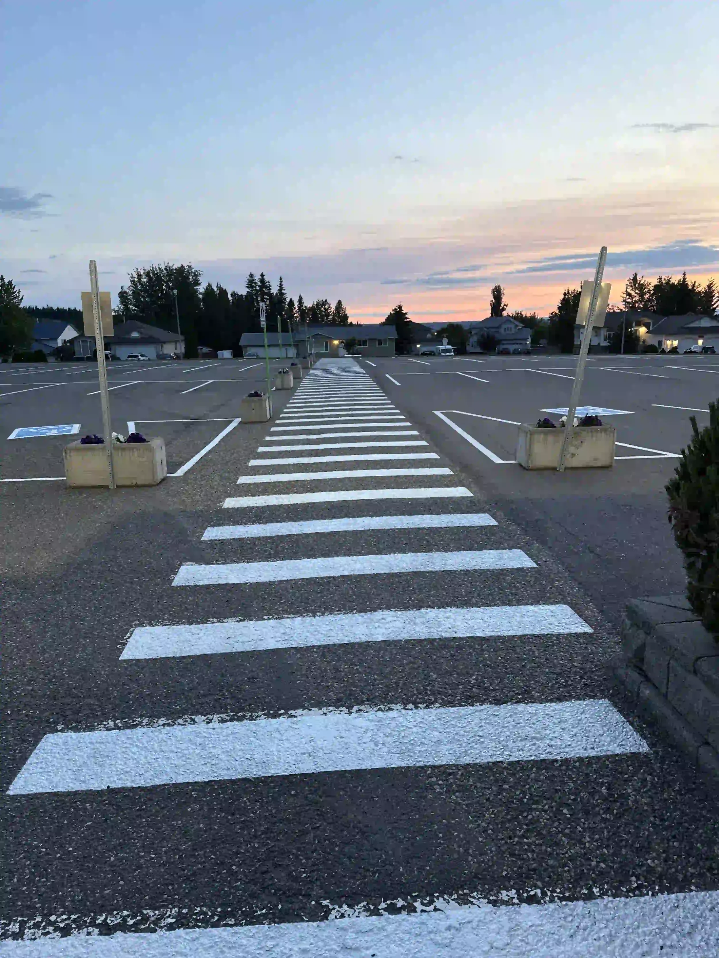 An empty parking lot during sunset with a pedestrian crosswalk and some sidewalk planters.