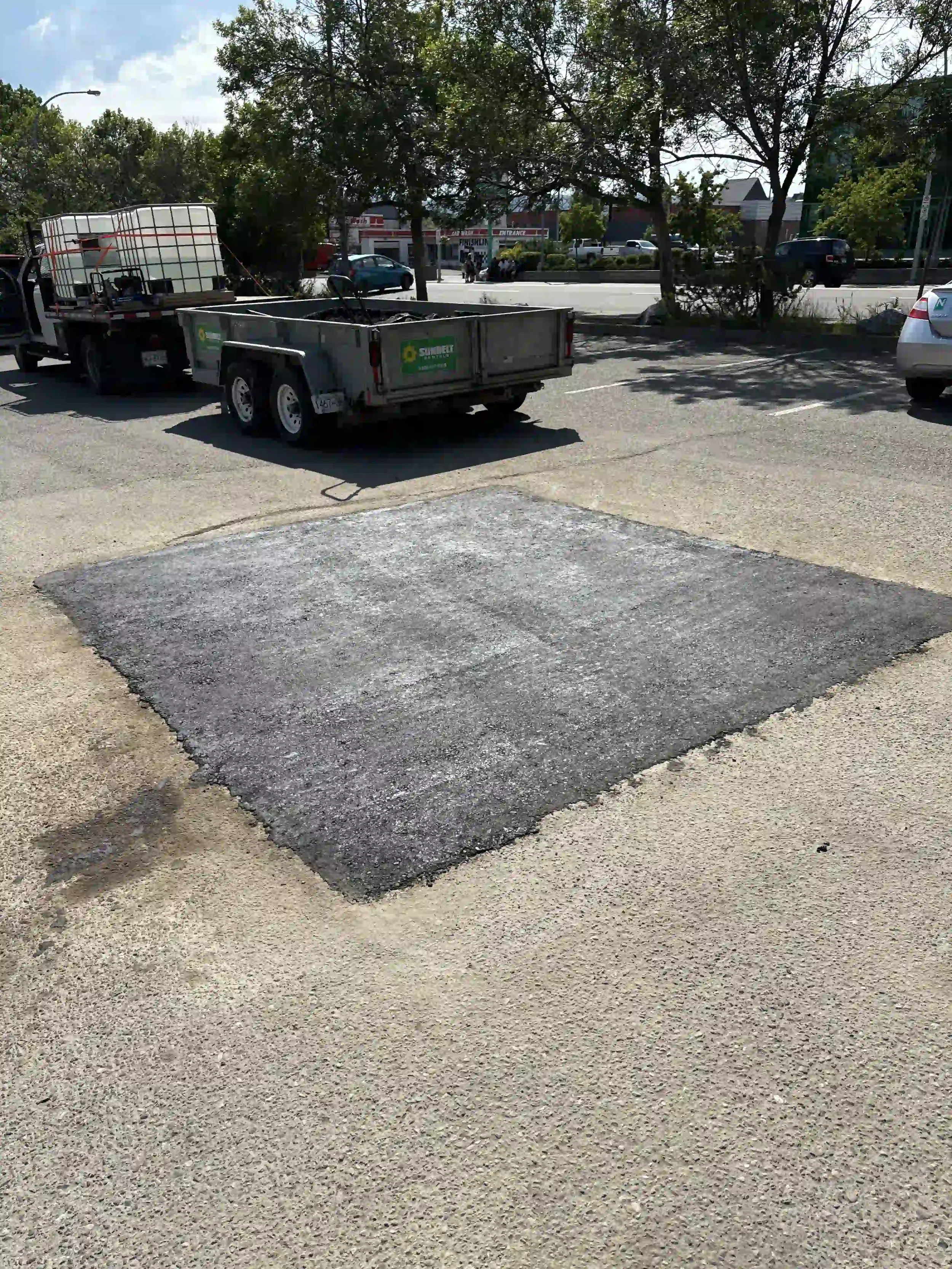 Empty parking lot with a large square patch of black asphalt, surrounded by gravel and concrete, with a trailer, a white car, and trees in the background.