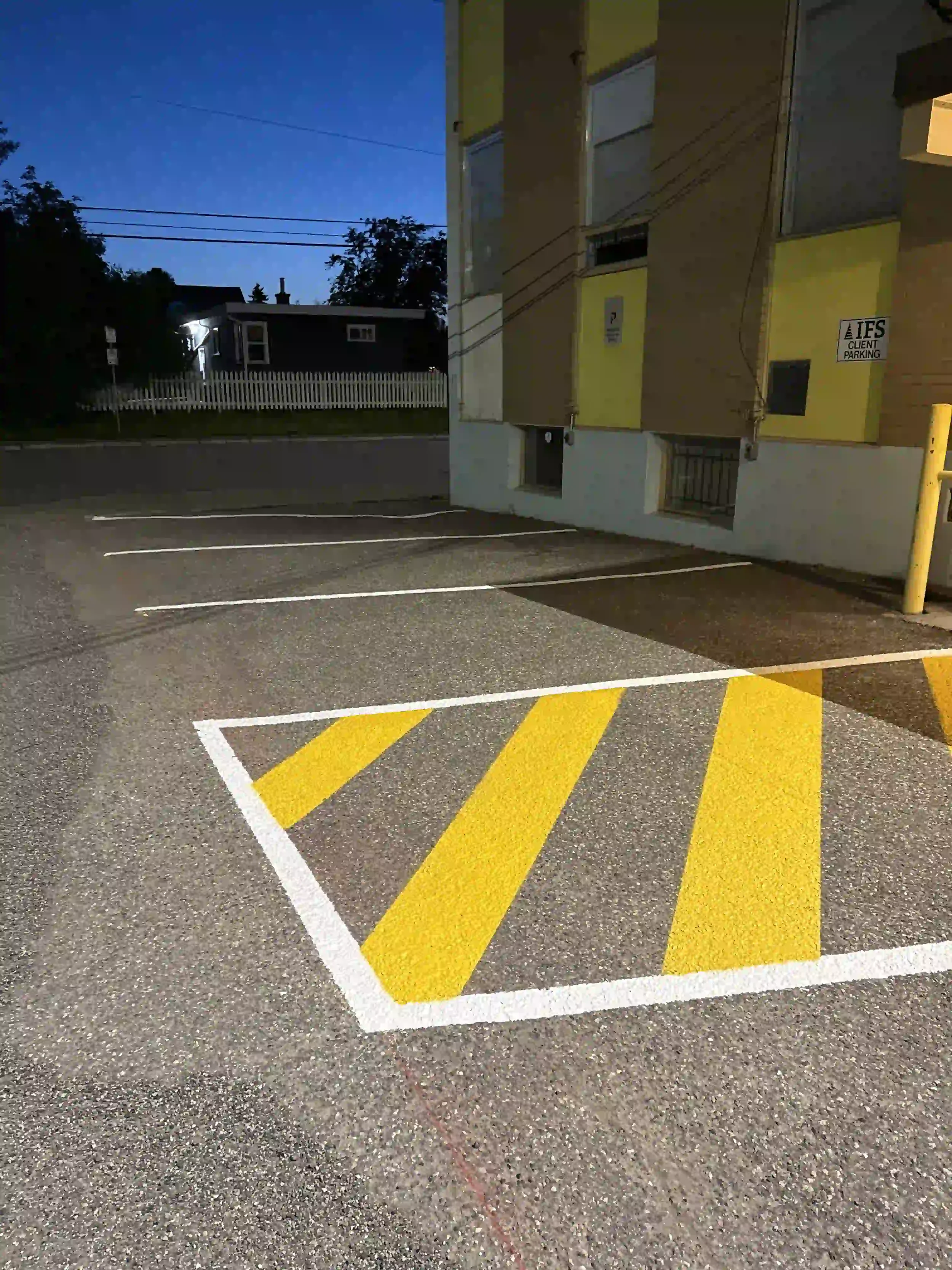Empty parking lot with marked parking spaces, yellow and white striped area in the foreground, building with yellow and brown exterior in the background, and twilight sky overhead.
