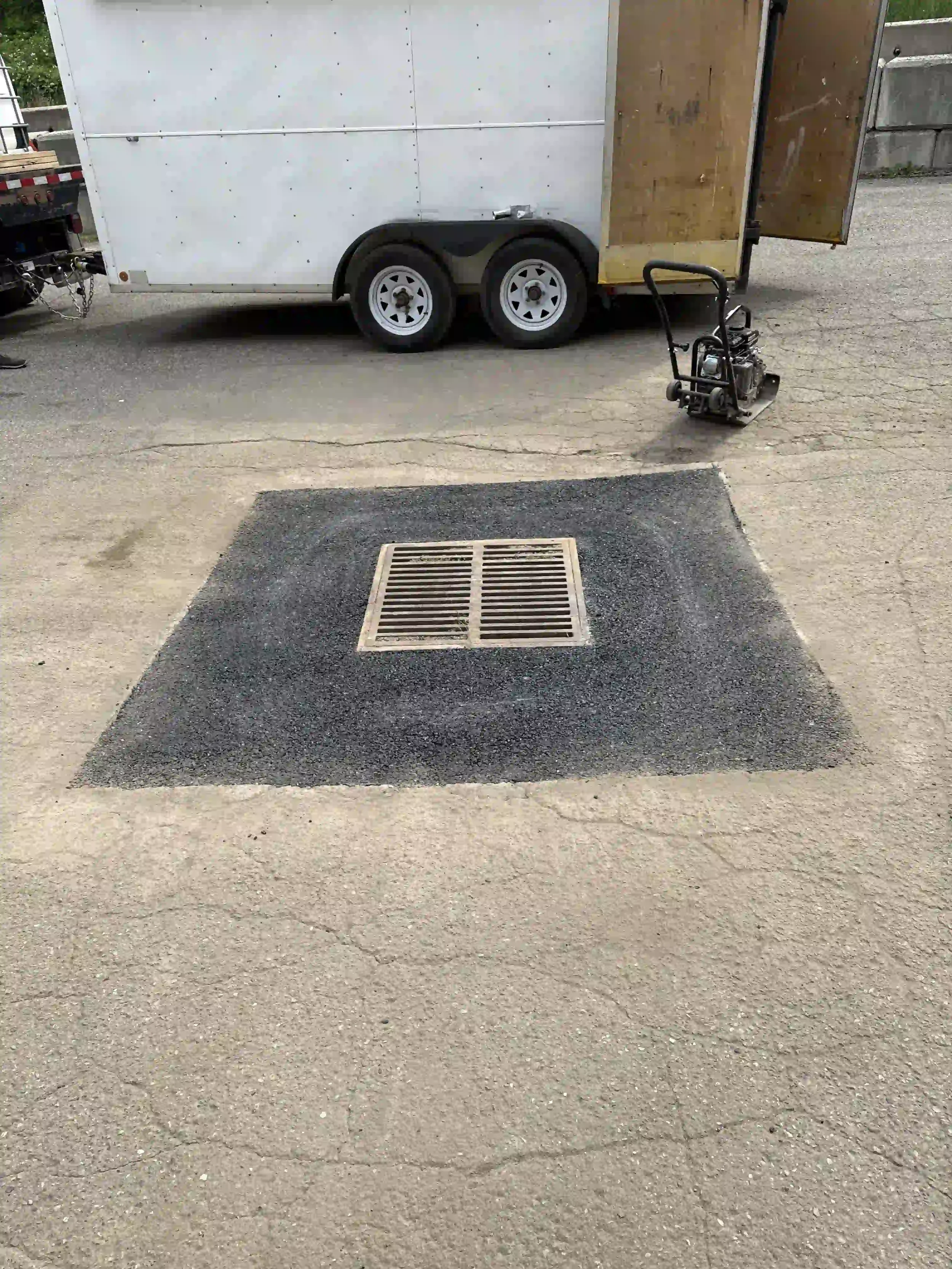 A street drainage grate embedded in a brown asphalt pavement, with a darker asphalt patch around it, on a gray concrete surface.