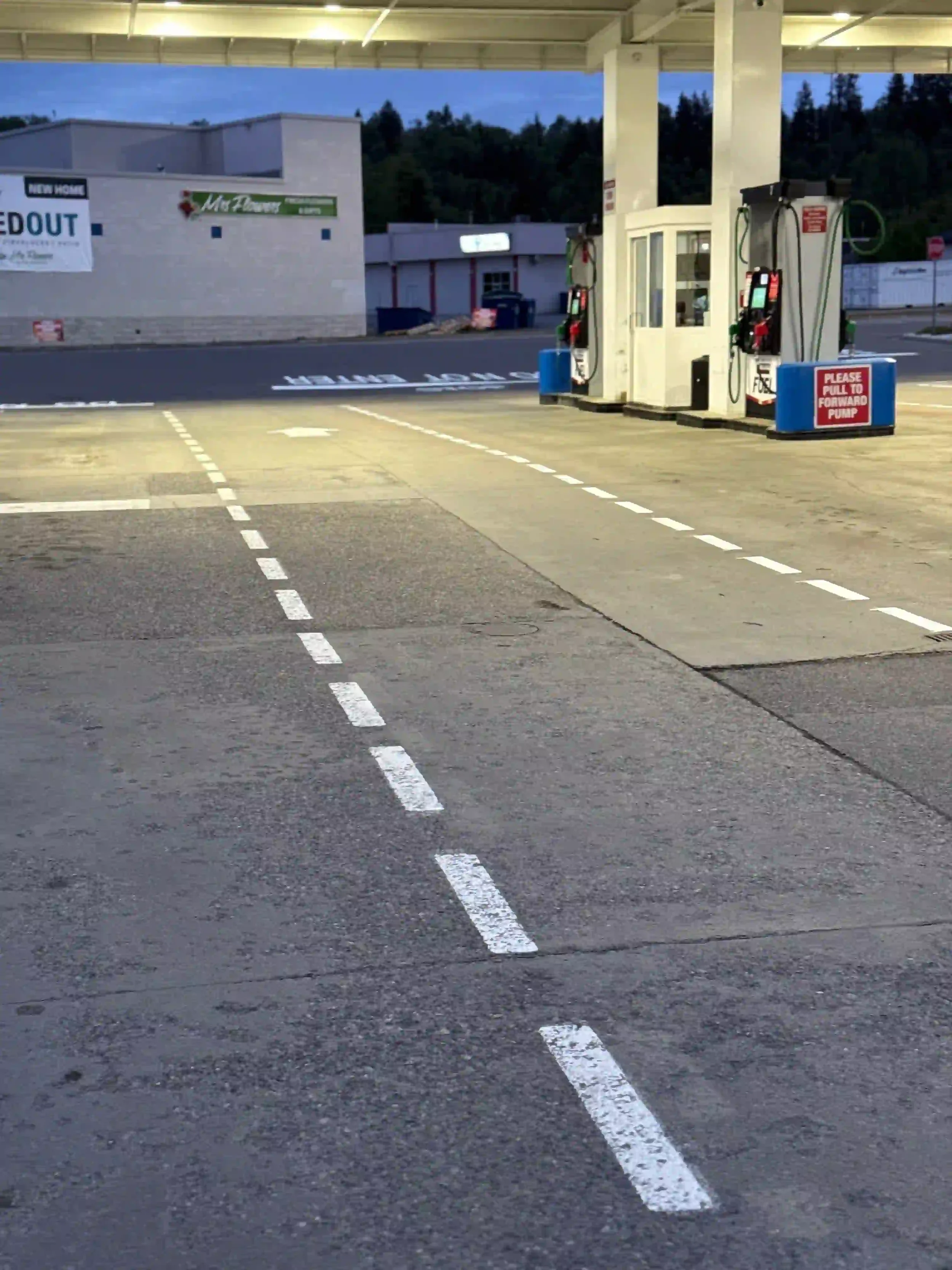 Empty gas station with two pumps and a small building in the background, with a sign that reads 'Please pull to forward pump' and a message about a new home auction on a banner.