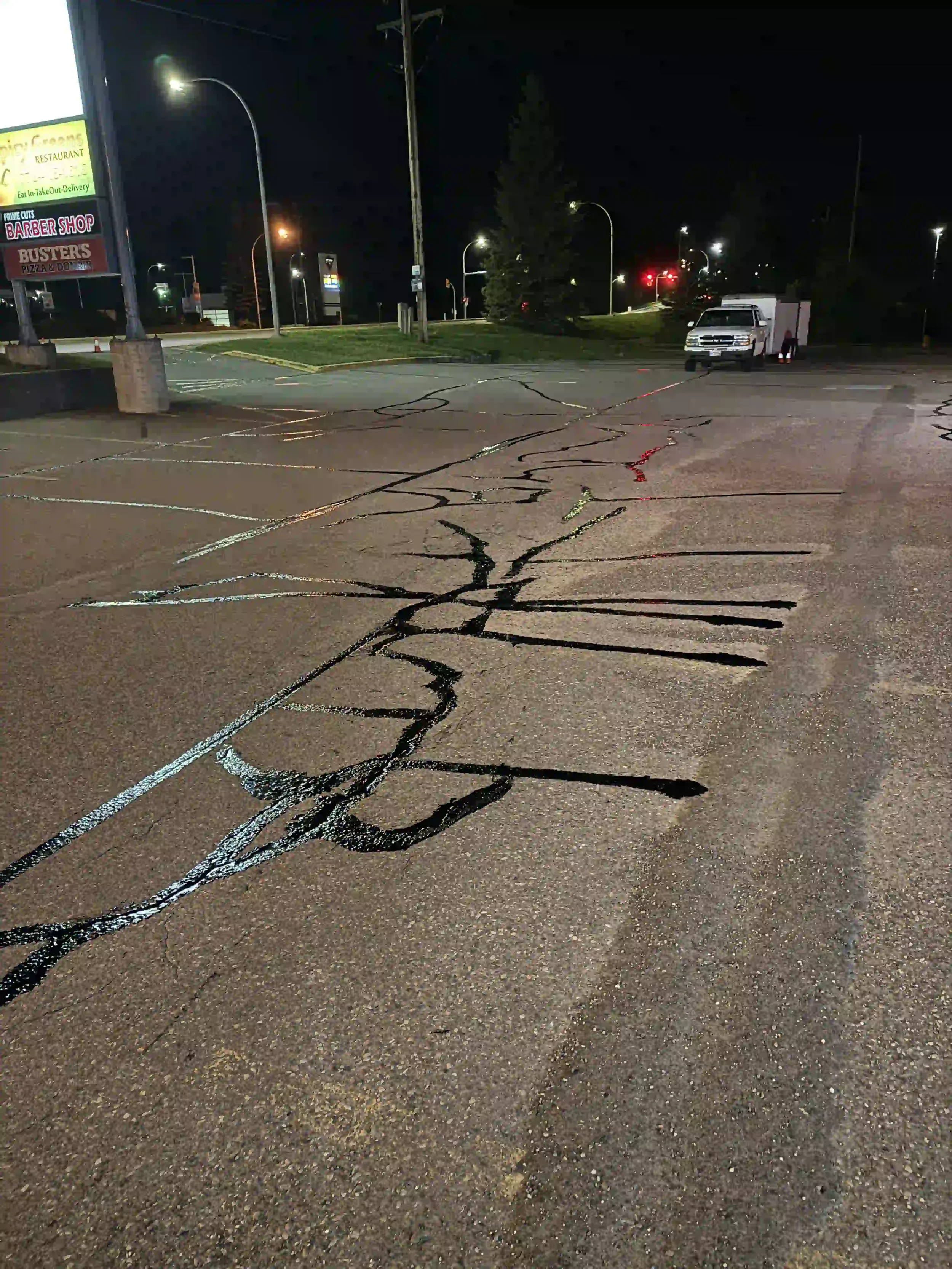 Nighttime view of a parking lot with visible cracks in the asphalt, a white utility truck parked in the distance, and illuminated commercial signs and streetlights.