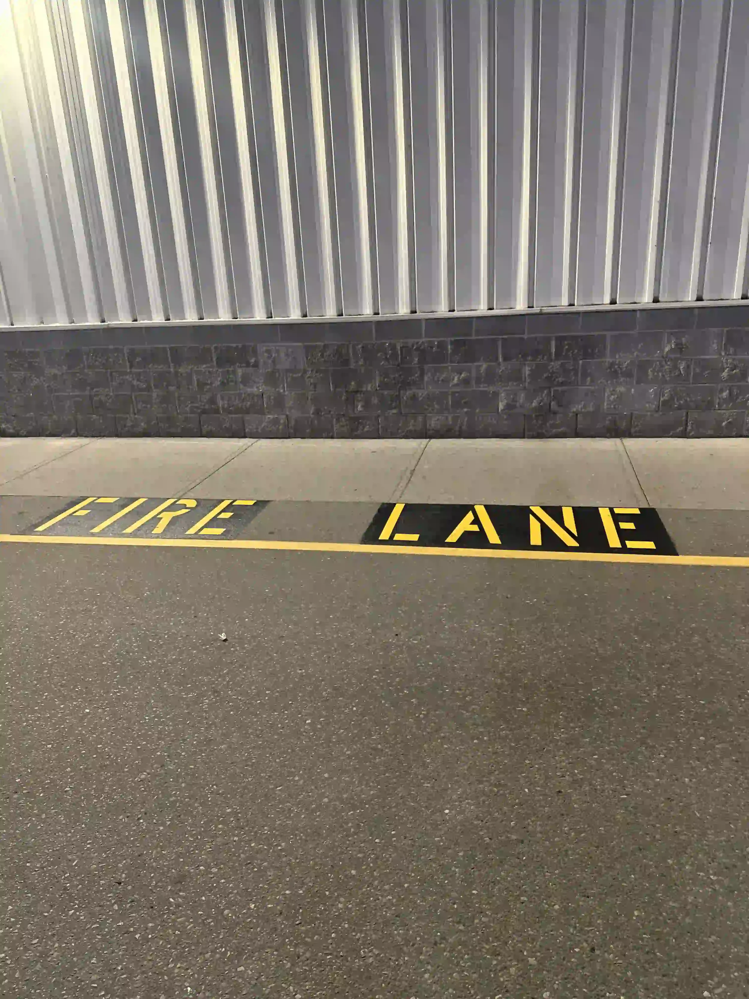 A parking lot ground with yellow painted words that read "FIRE LANE" and a metal wall with vertical ridges in the background.