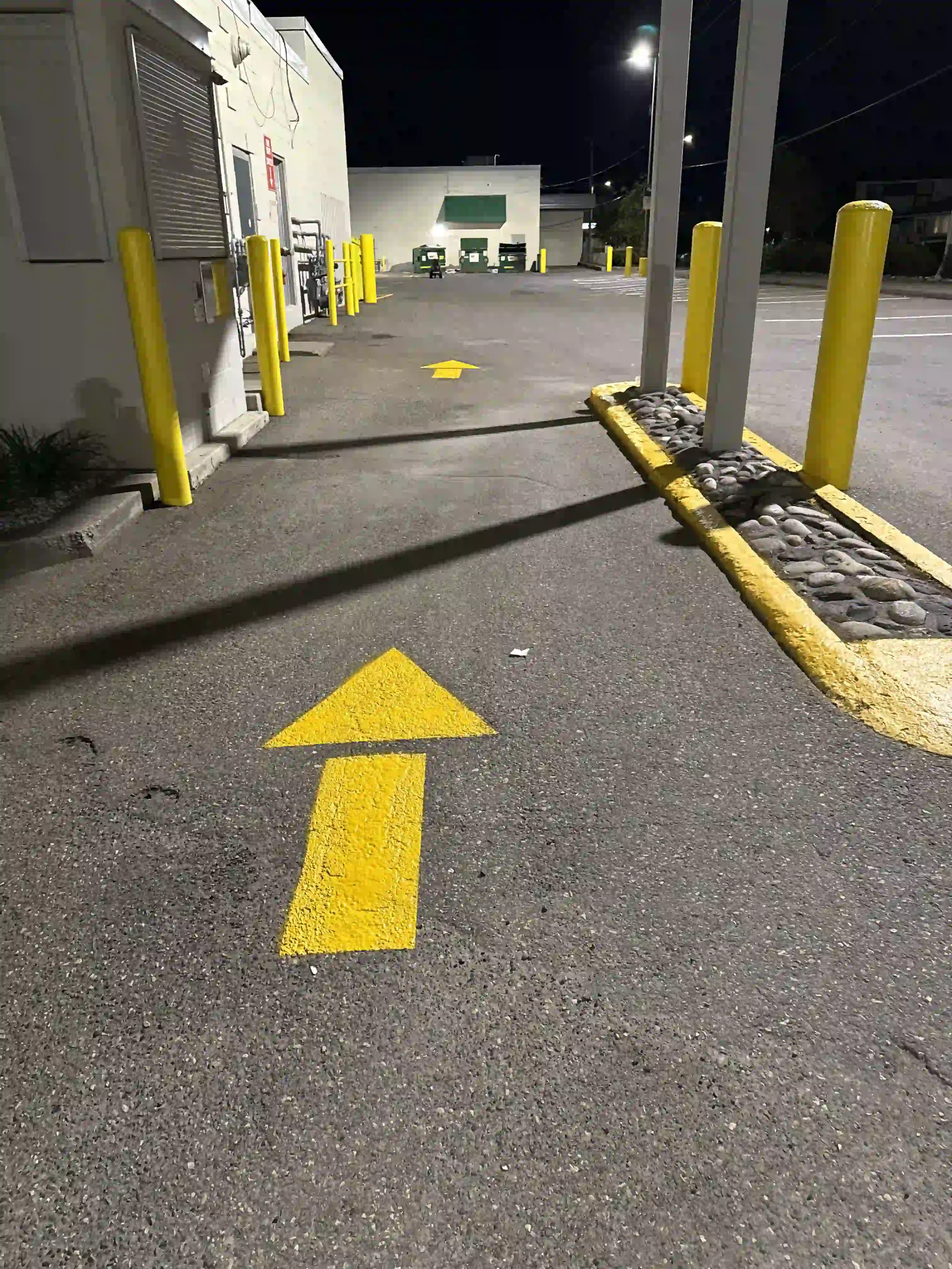 Nighttime view of an empty parking lot with yellow directional arrows painted on the asphalt, yellow safety posts, and a row of dumpsters in the background. A building with a green awning and a large vent on the wall is visible in the distance.