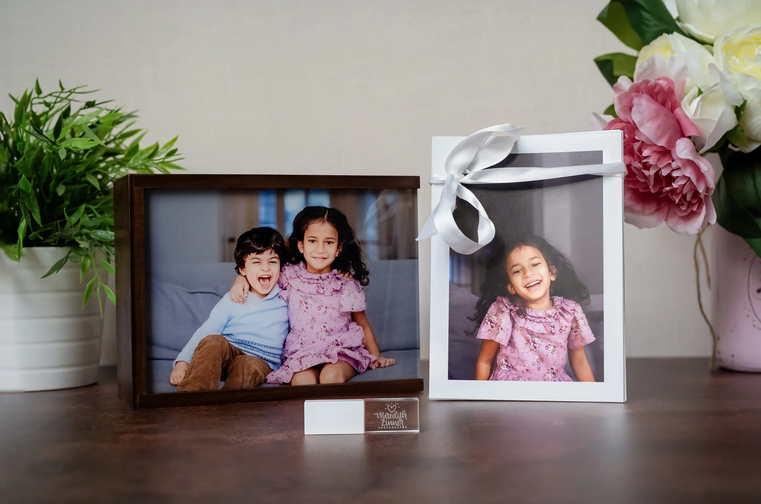 A framed photo of two children, a boy and a girl, sitting together and smiling, with a larger printed picture of the girl beside it, surrounded by a green potted plant and pink flowers.