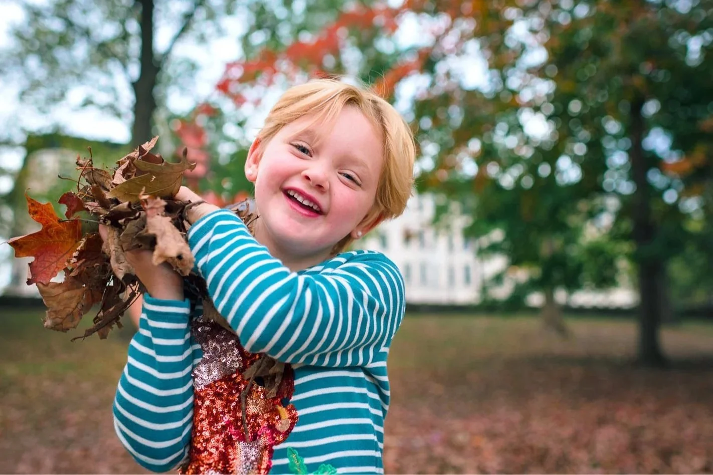 A young girl with red hair and a striped teal and white long-sleeve shirt smiling and holding a bunch of fallen autumn leaves outdoors in a park with trees in the background.