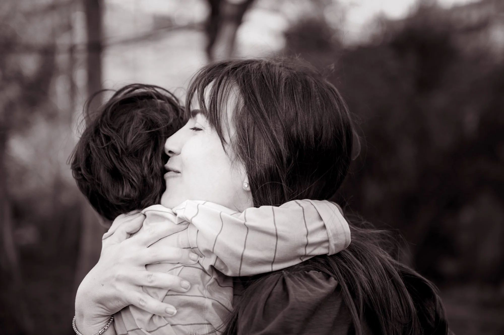 A woman with long dark hair hugging a child with short curly hair. The woman is smiling with closed eyes as she embraces the child, and the background is blurred with trees.