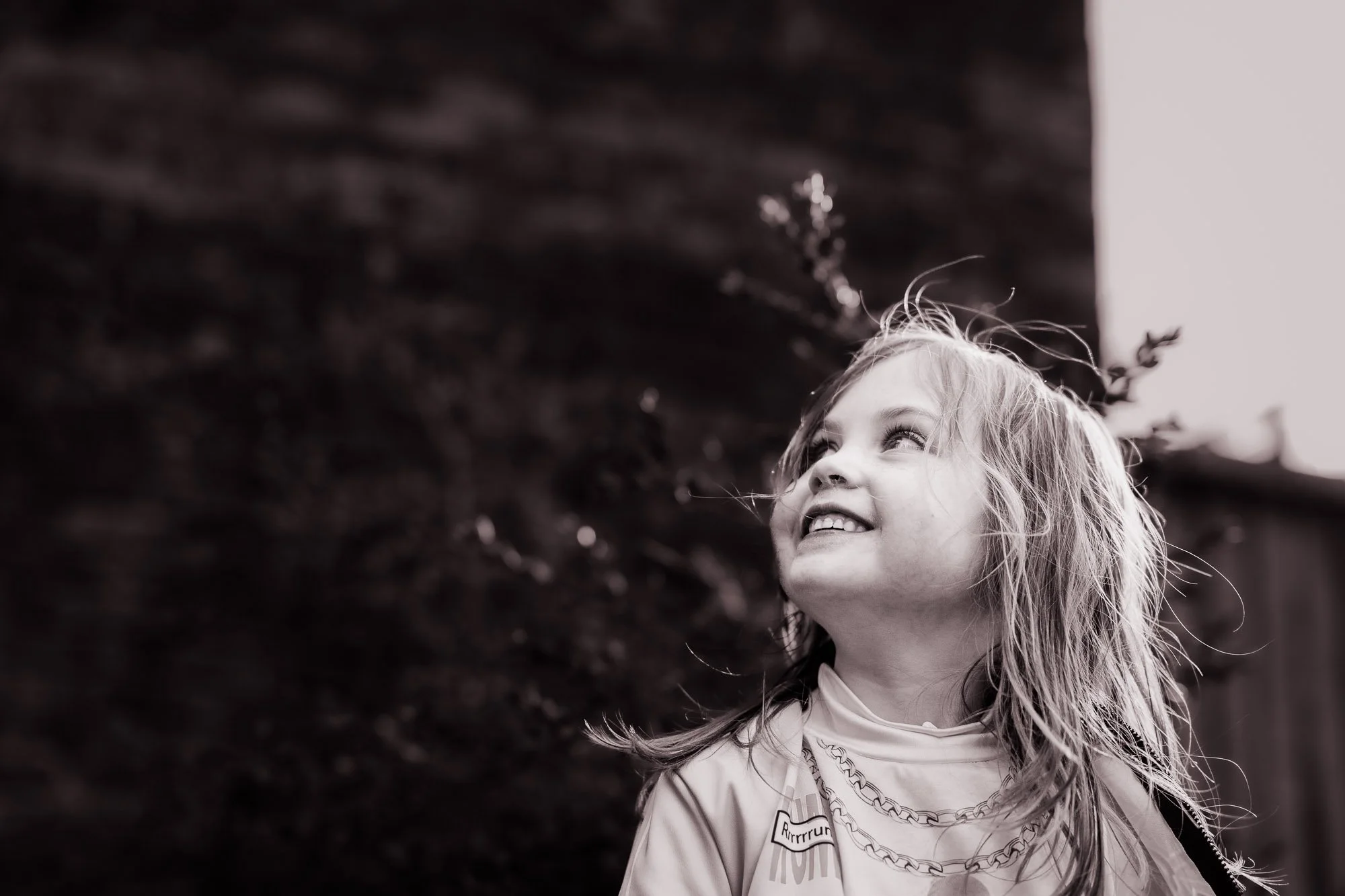 A young girl with long, curly hair smiling and looking upward outdoors, blurred dark foliage in the background, black and white photo.