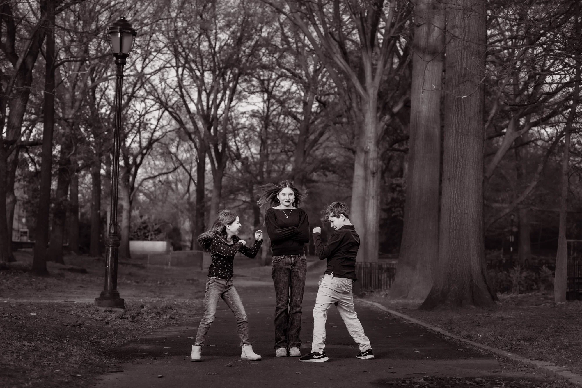 Four kids playing and talking in a park with tall trees and a street lamp on a cloudy day, in black and white.