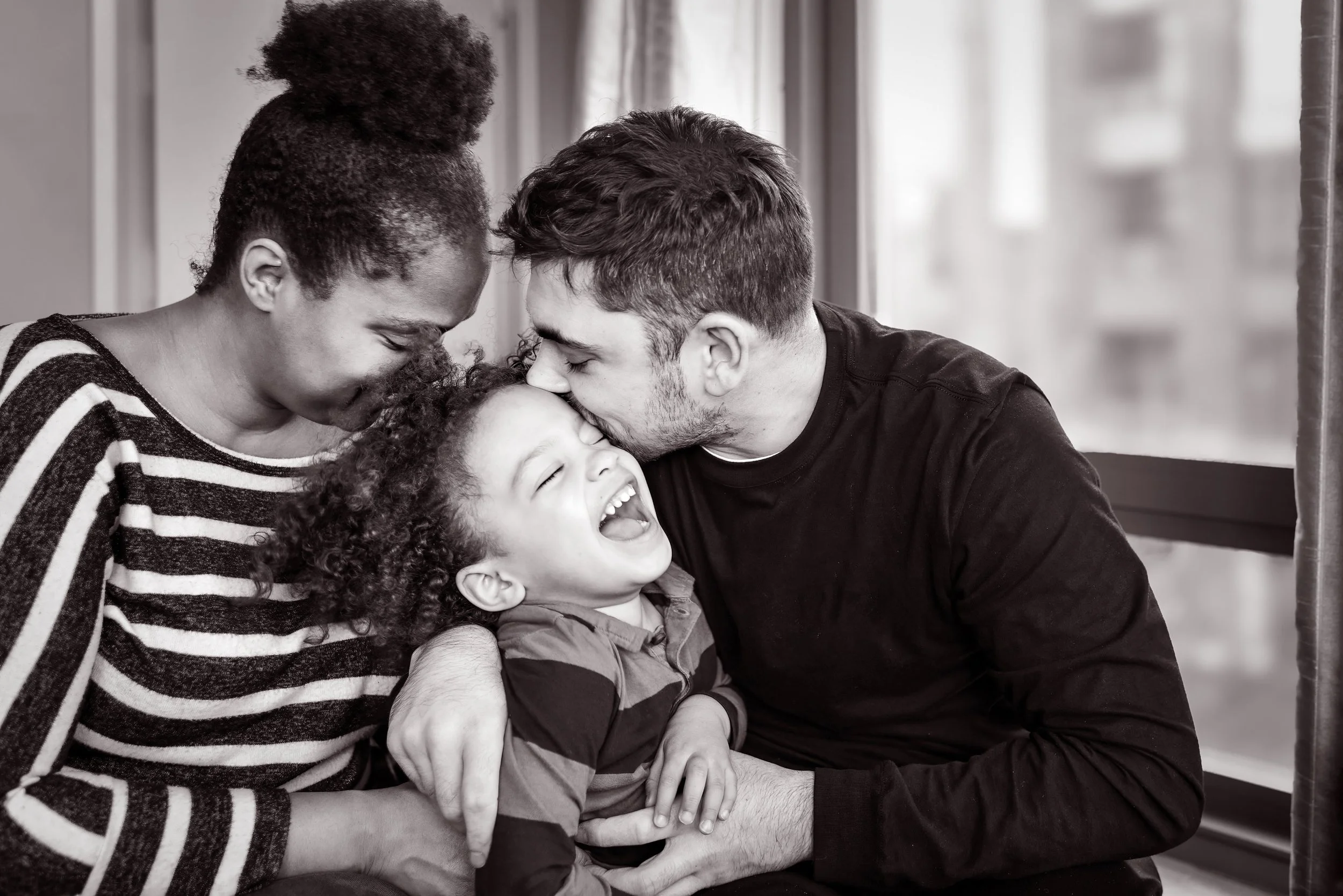 A playful moment showing a woman, man, and young girl laughing as they sit closely together with their foreheads touching inside a room with curtains and windows.