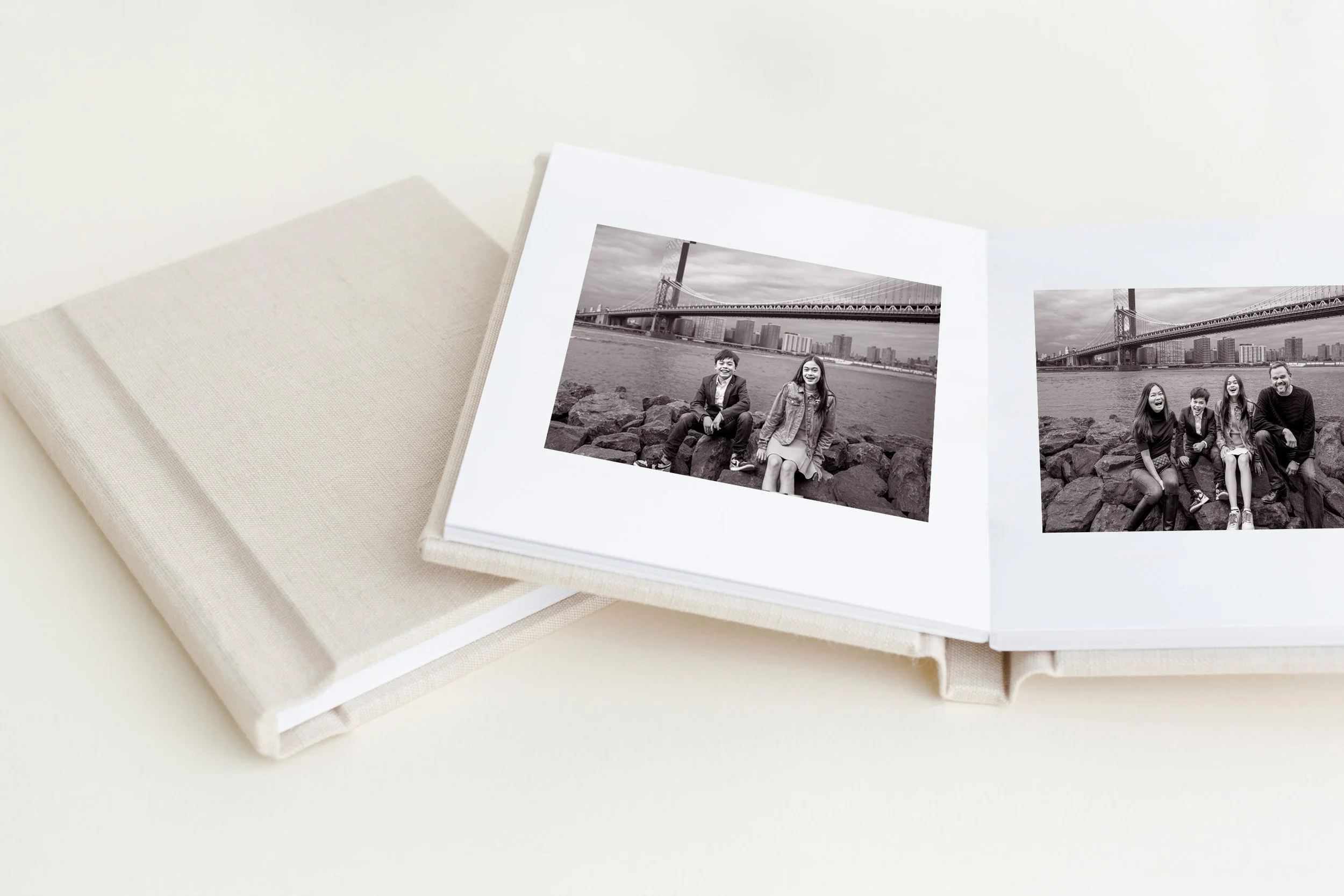 Open photo album with black and white pictures of people in front of bridge and city skyline, on white fabric-covered surface