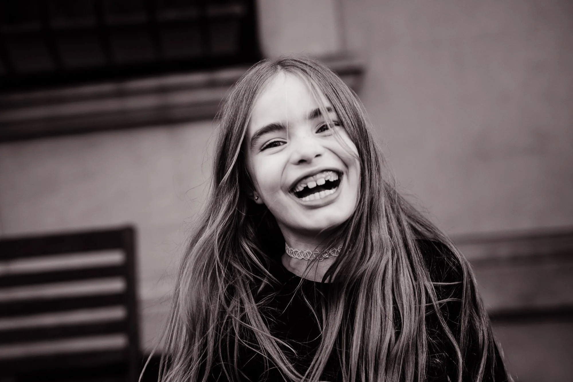 A young girl with long hair laughing and showing her teeth, wearing a choker necklace and large earrings, with a blurred background.