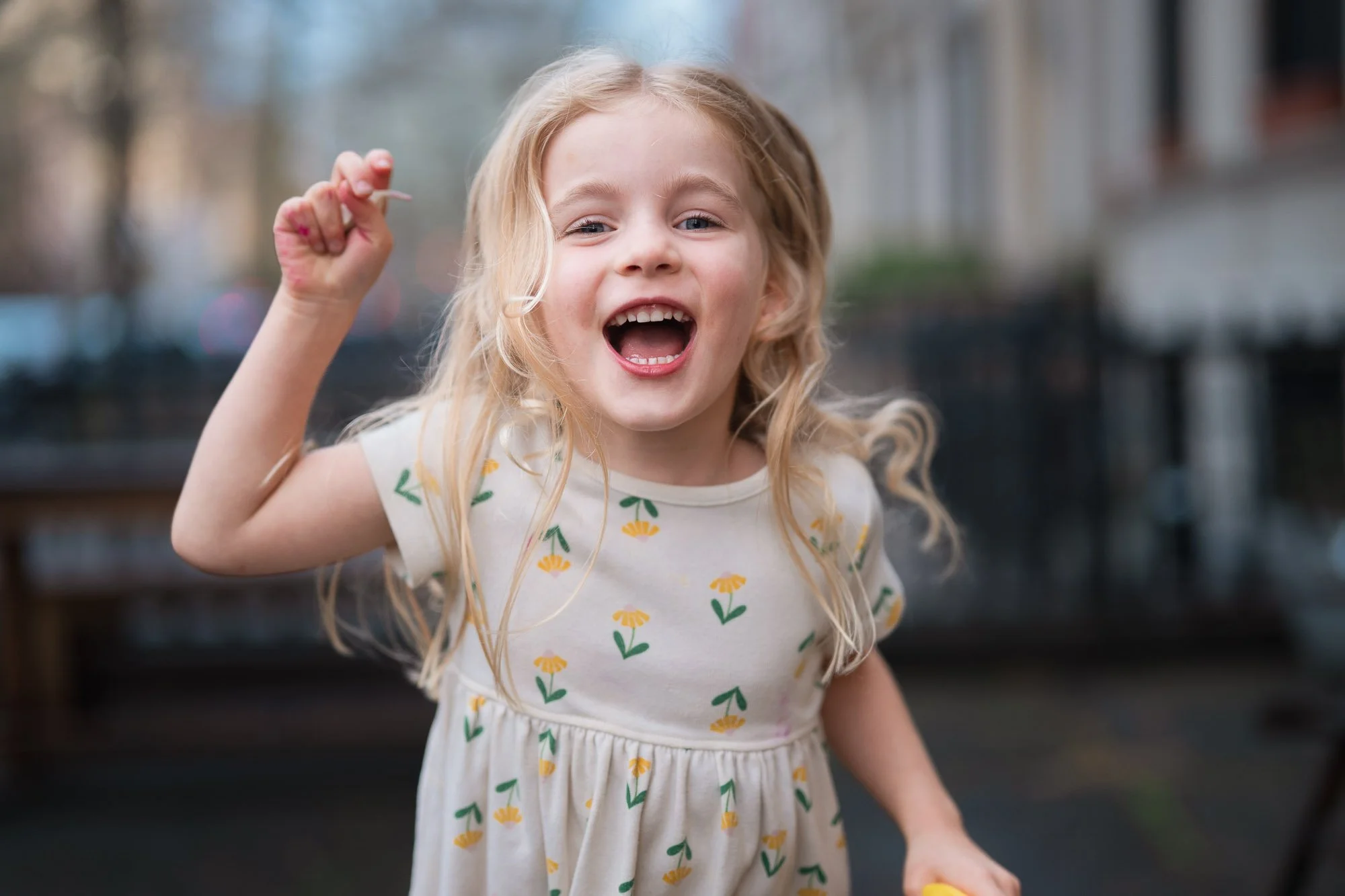 A young girl with blonde hair, wearing a white dress with yellow and green floral patterns, is smiling excitedly and holding a small object in her hand outdoors.