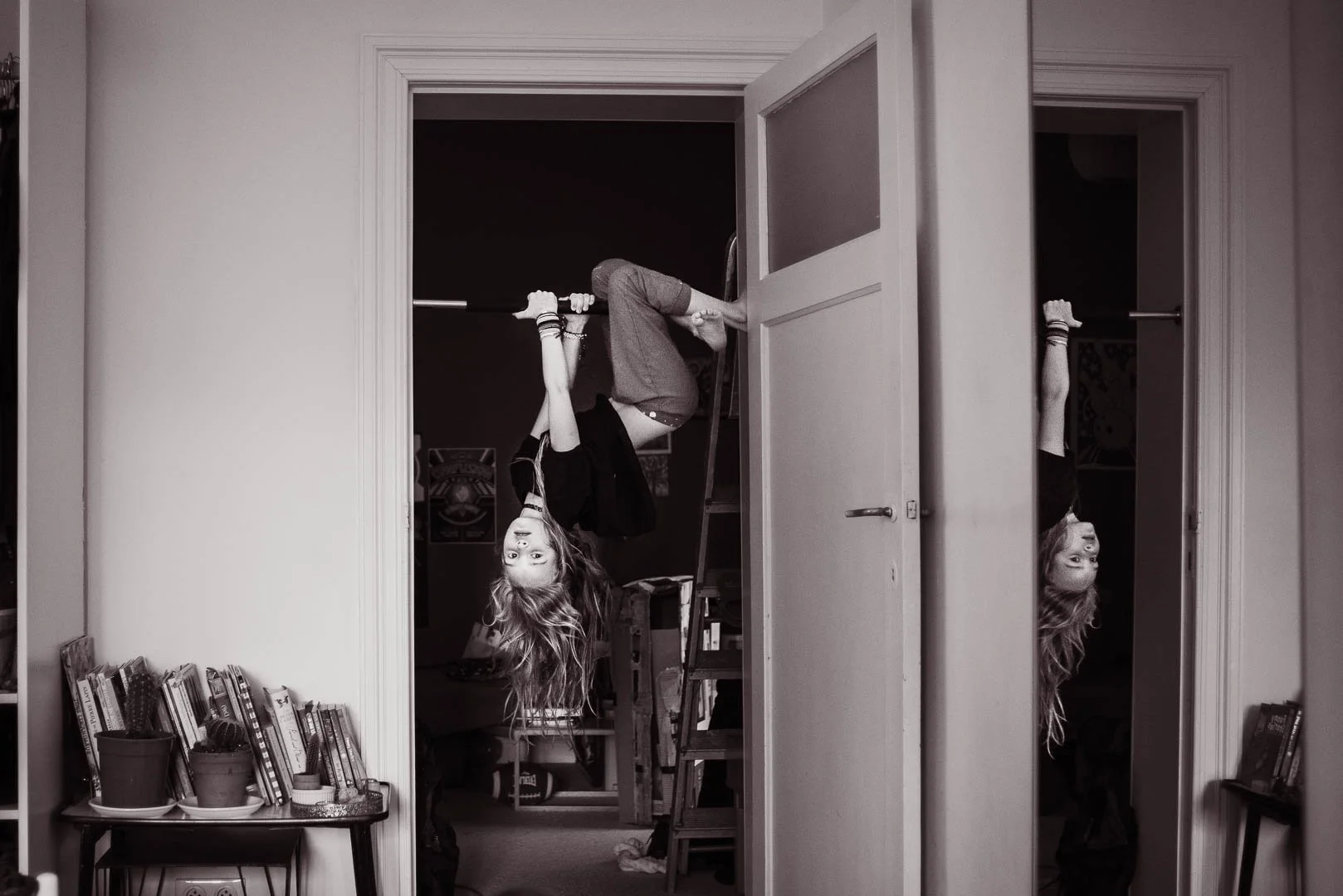 A girl hanging upside down from a pole with her legs over a doorway, her head and hair hanging towards the floor, seen in a reflection in a mirror on the right side of the image.